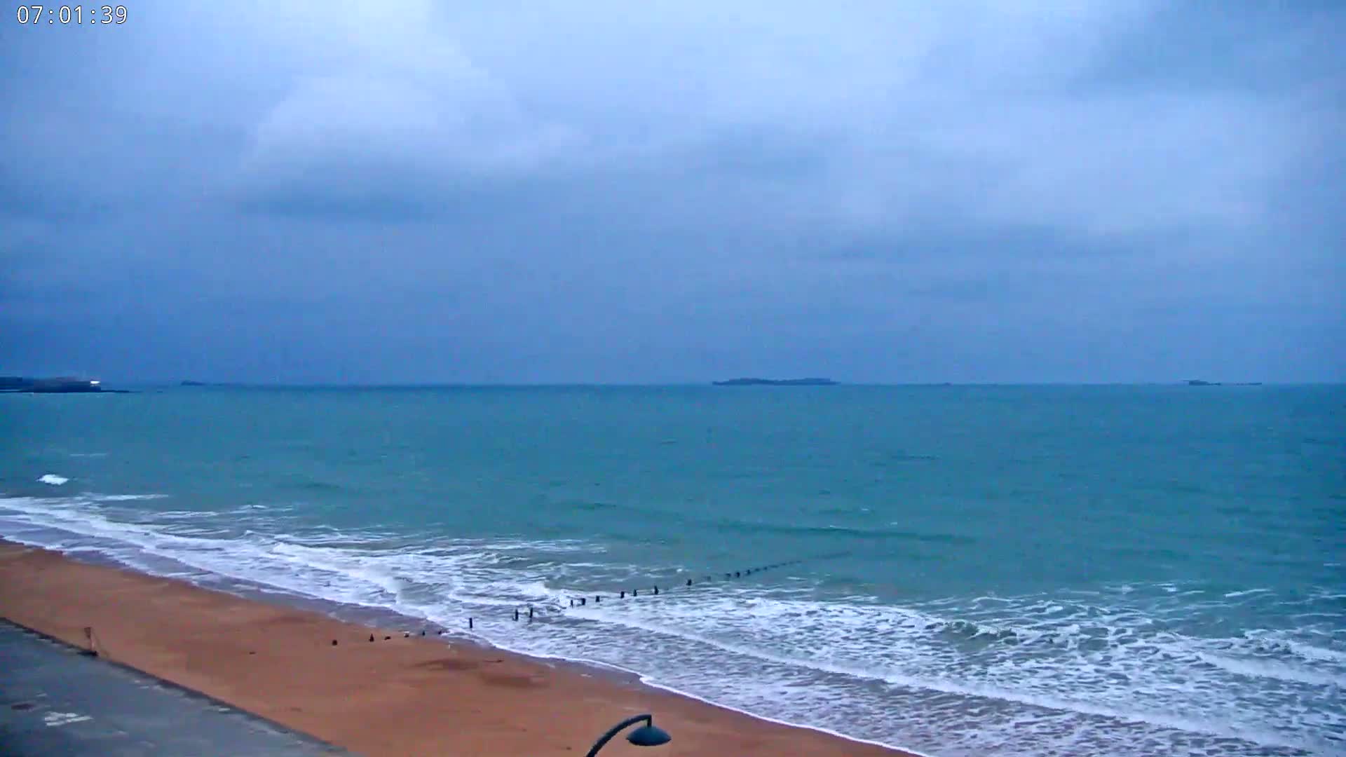 A sandy beach with gentle waves rolls onto the shore under a cloudy sky.
