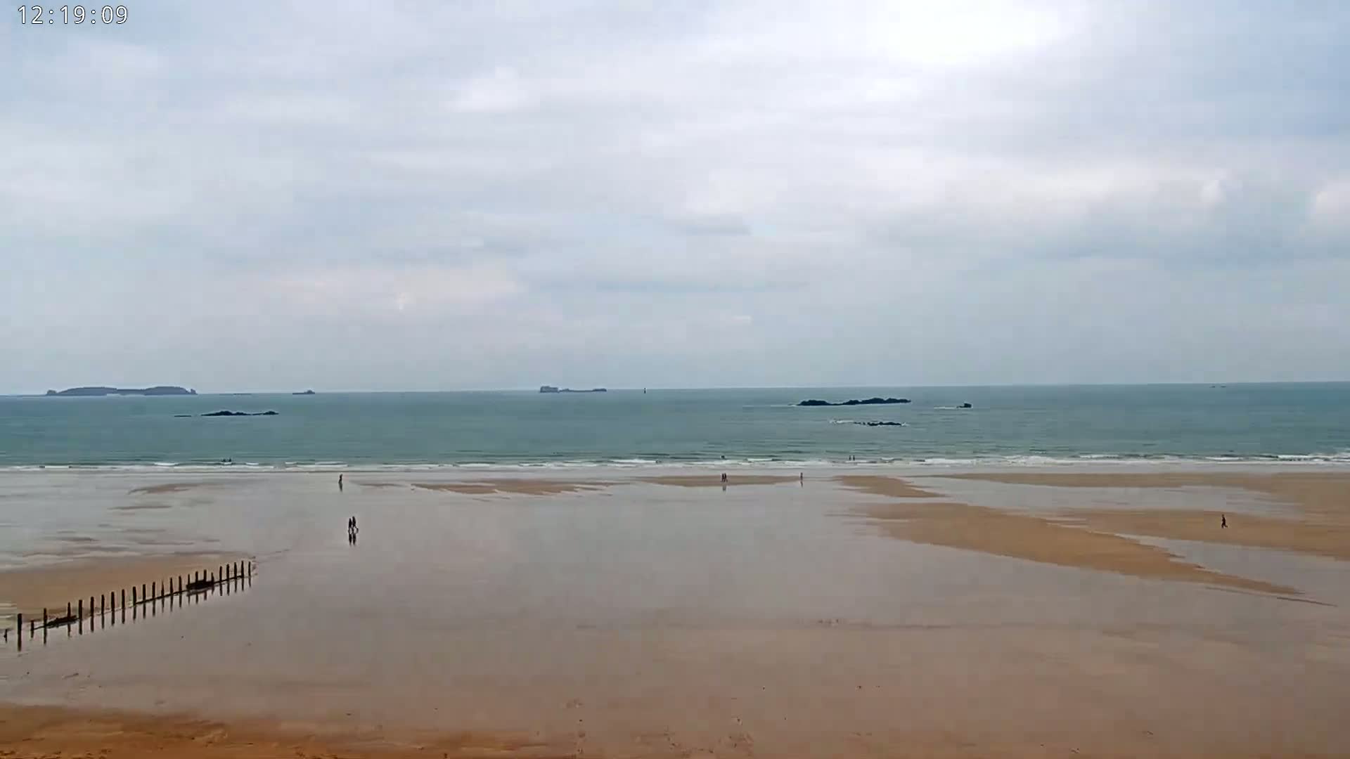 A wide sandy beach stretches out to a calm, teal ocean under a cloudy sky, with a few people scattered along the shore and some distant islands visible on the horizon.