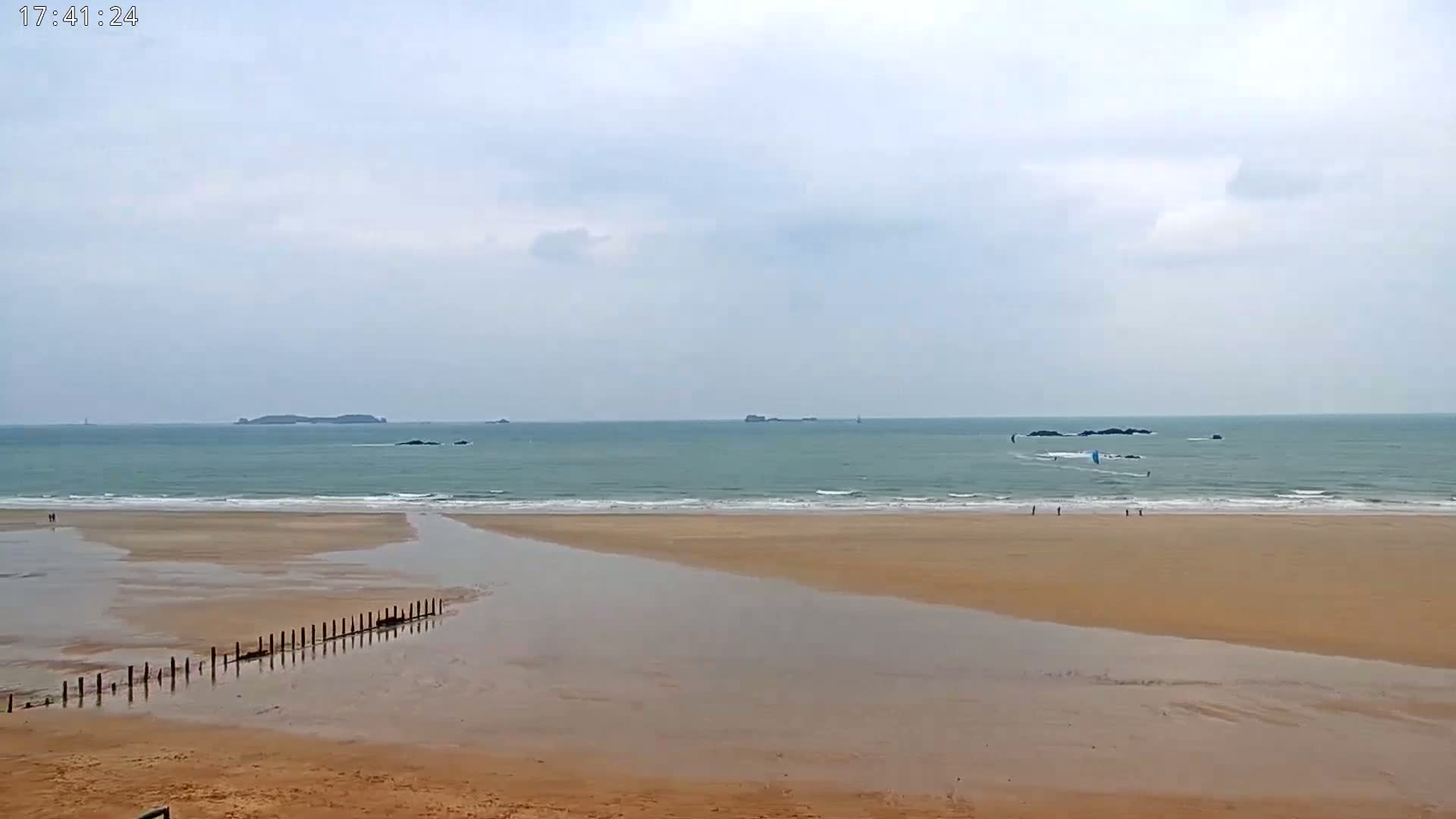 A wide sandy beach with a calm ocean under an overcast sky, featuring a few people and windsurfers in the distance.