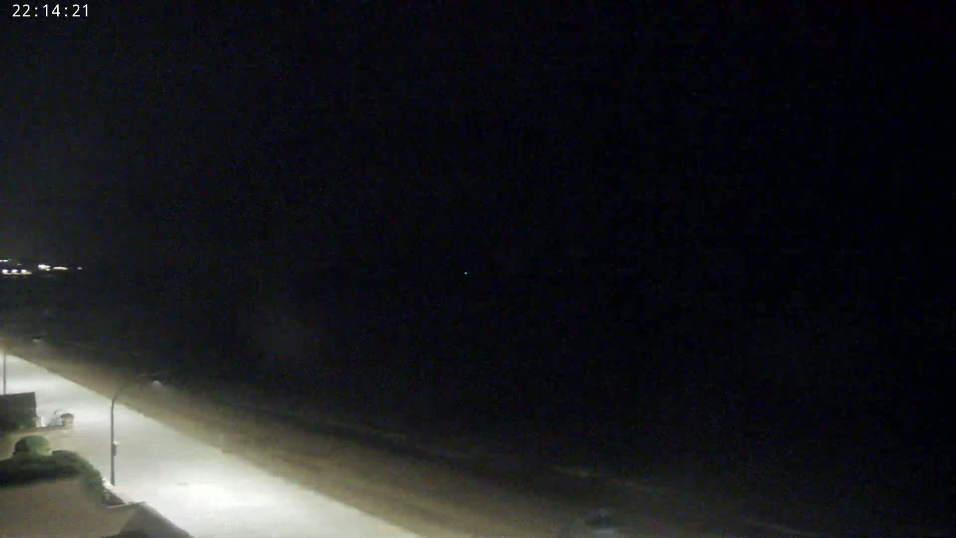 A nighttime view of a dark beach with a paved walkway lit by streetlights under a clear, starlit sky.
