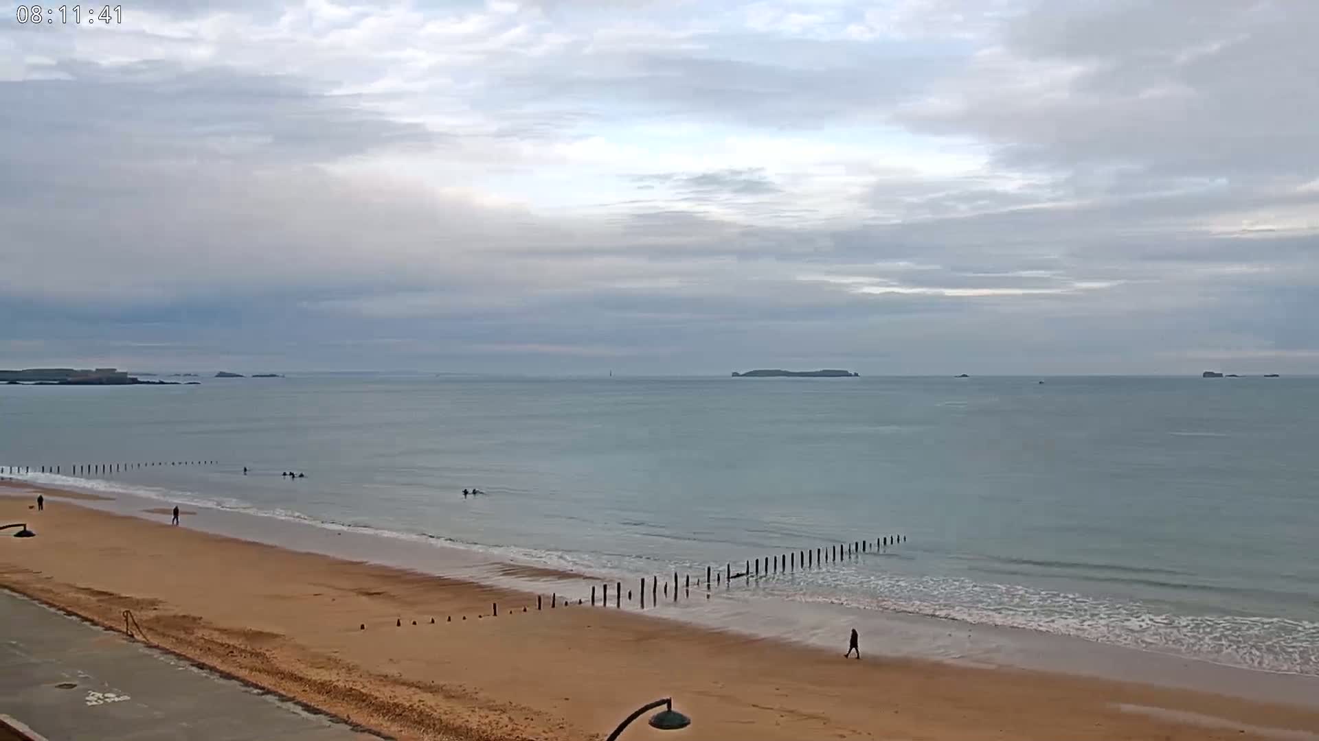 A sandy beach stretches toward a calm sea under a cloudy sky, with a few people walking and swimming.