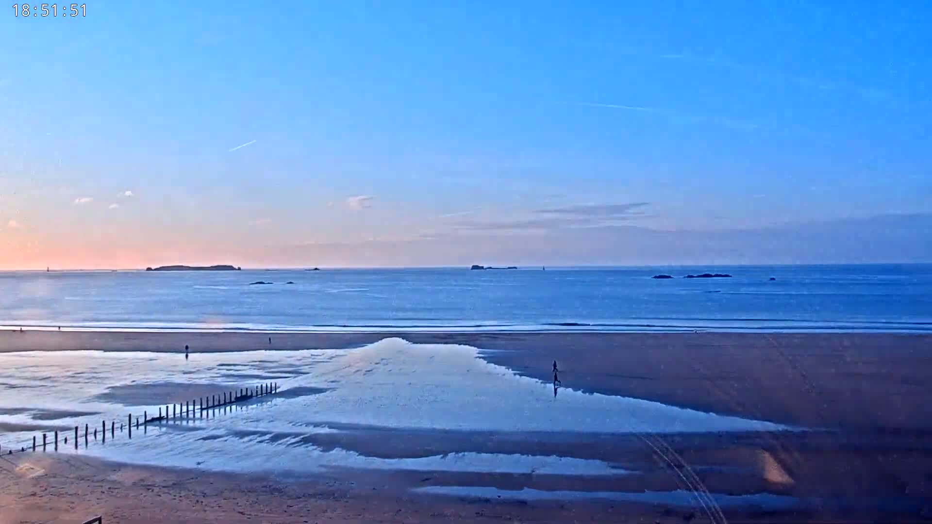 A sandy beach at sunset, with a calm ocean and distant islands under a clear, pale blue sky.