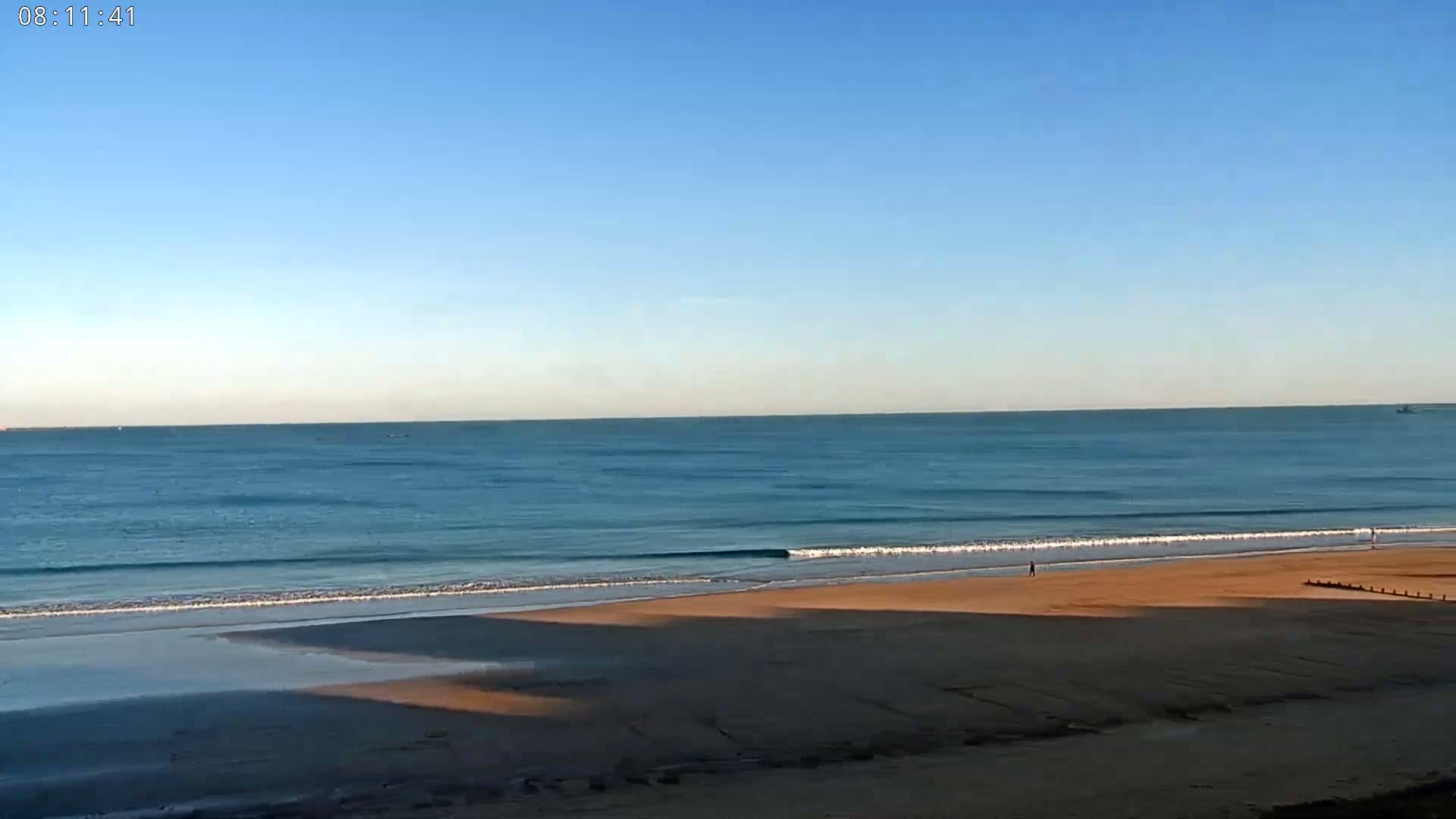 A calm ocean with small waves laps a sandy beach under a clear blue sky.