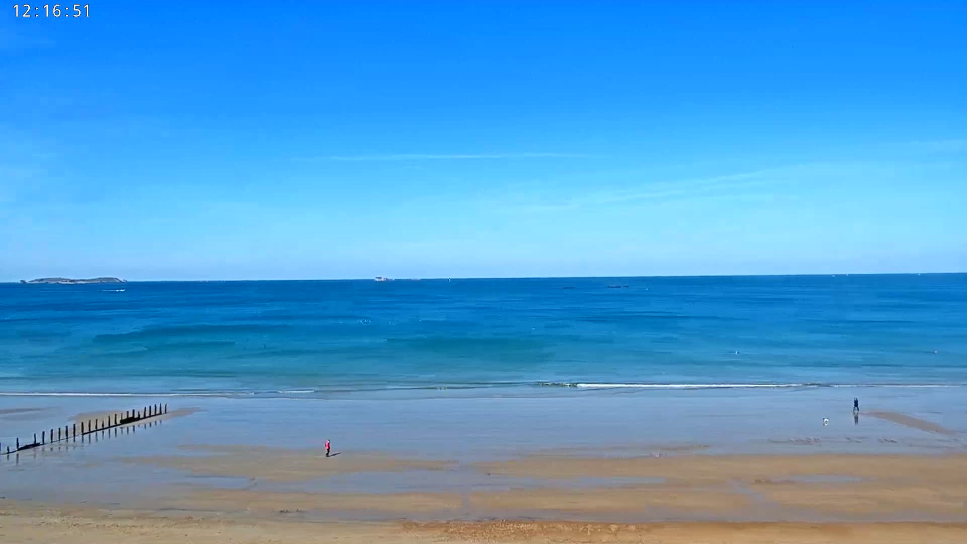 A sandy beach stretches to a calm, blue ocean under a clear blue sky, with a few people walking on the beach and a distant island visible on the horizon.