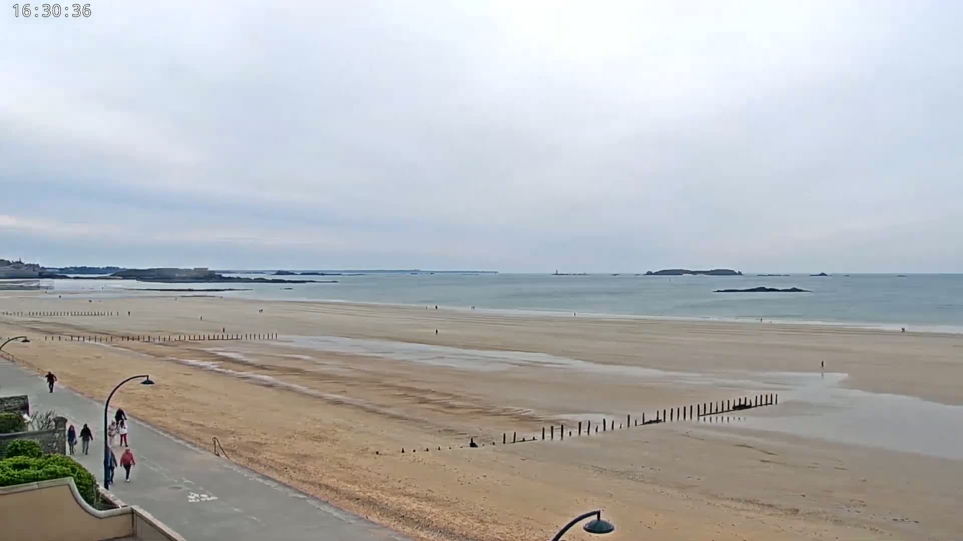 A wide sandy beach stretches to a calm ocean under an overcast sky, with a few people walking on the beach and along a paved path.