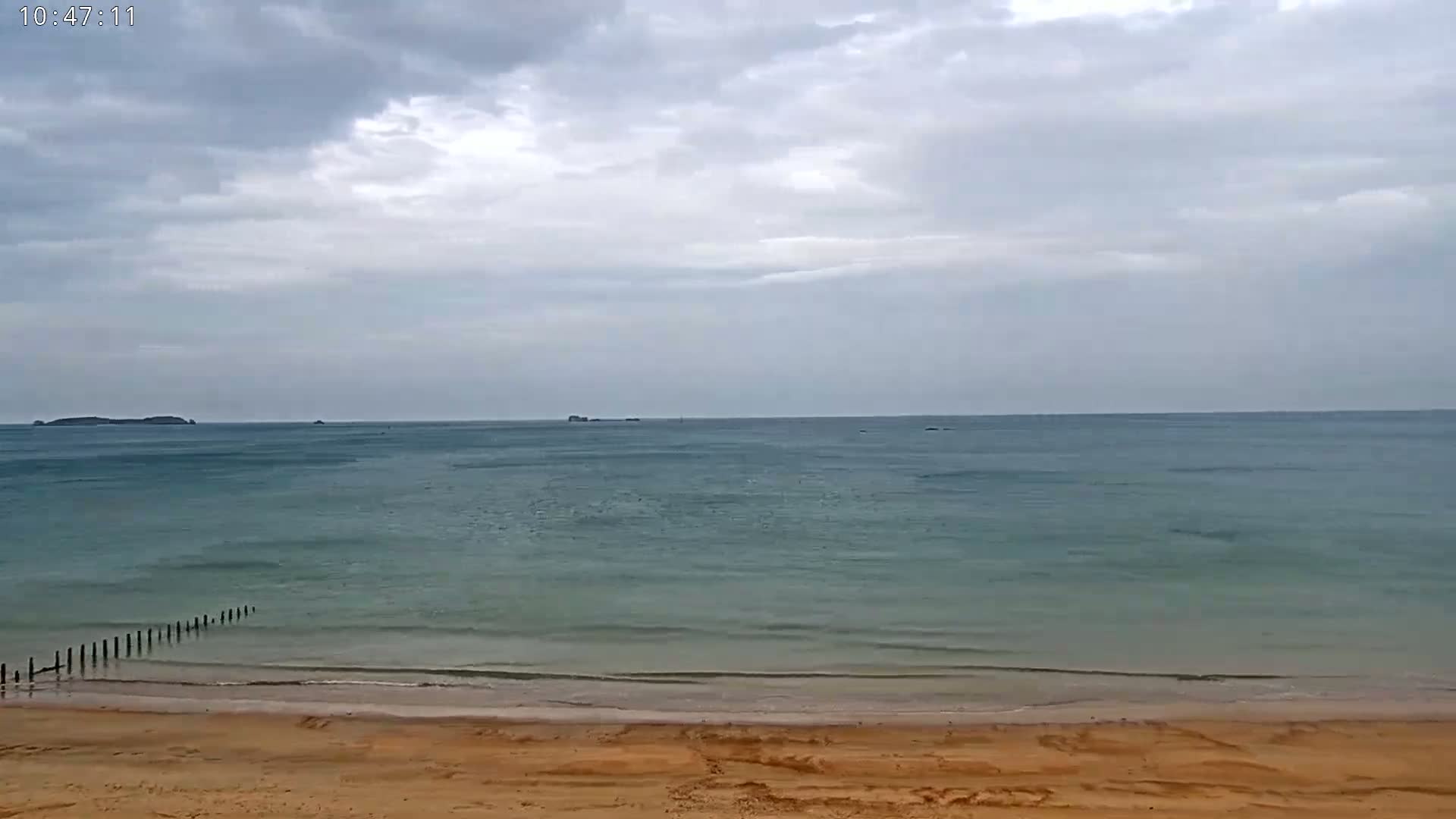 A sandy beach meets a calm, teal ocean under a cloudy sky, with distant islands visible on the horizon.