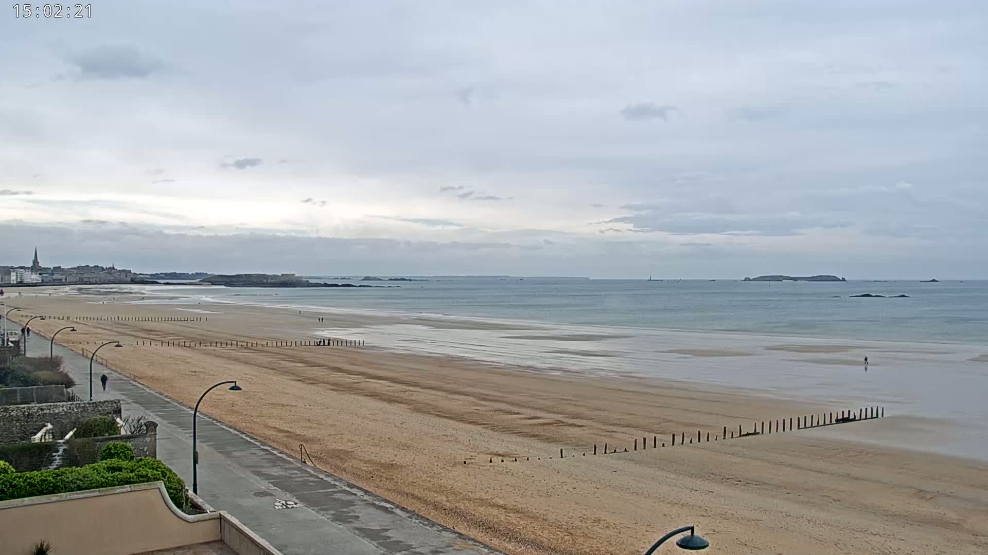 A long, sandy beach stretches out to a calm ocean under a cloudy sky, with a paved walkway and buildings visible in the foreground.