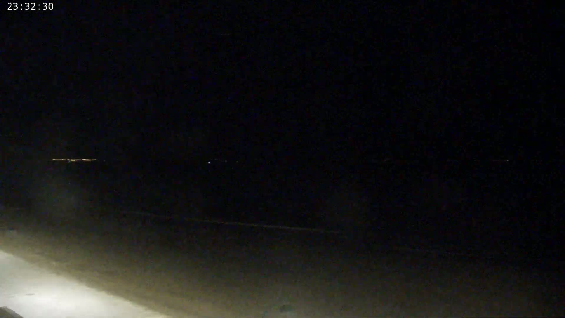 A dark, nighttime beach scene shows a faint horizon line with distant lights, under a clear, black sky.