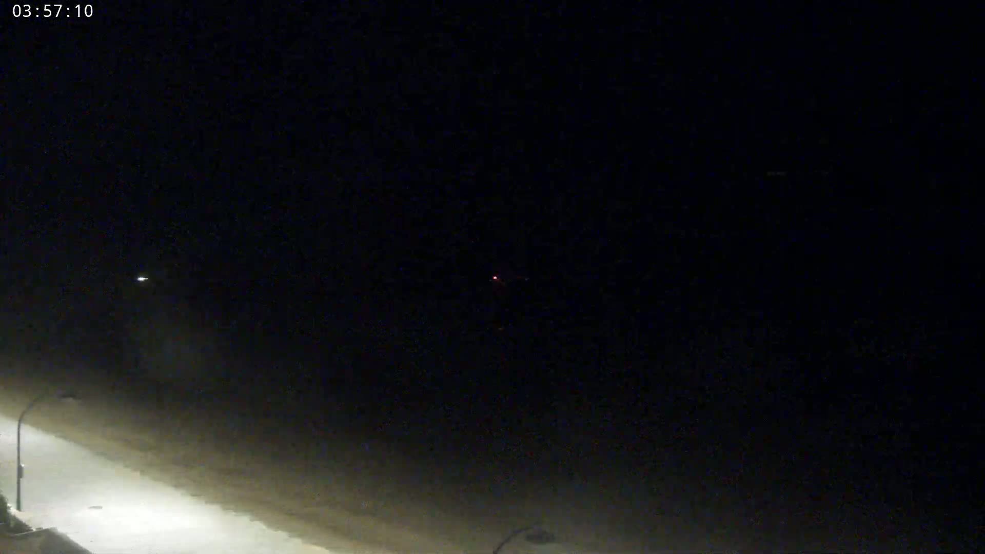 A nighttime, low-angle view shows a dark road illuminated by streetlights, extending towards a dark sky with a faint light source in the distance.