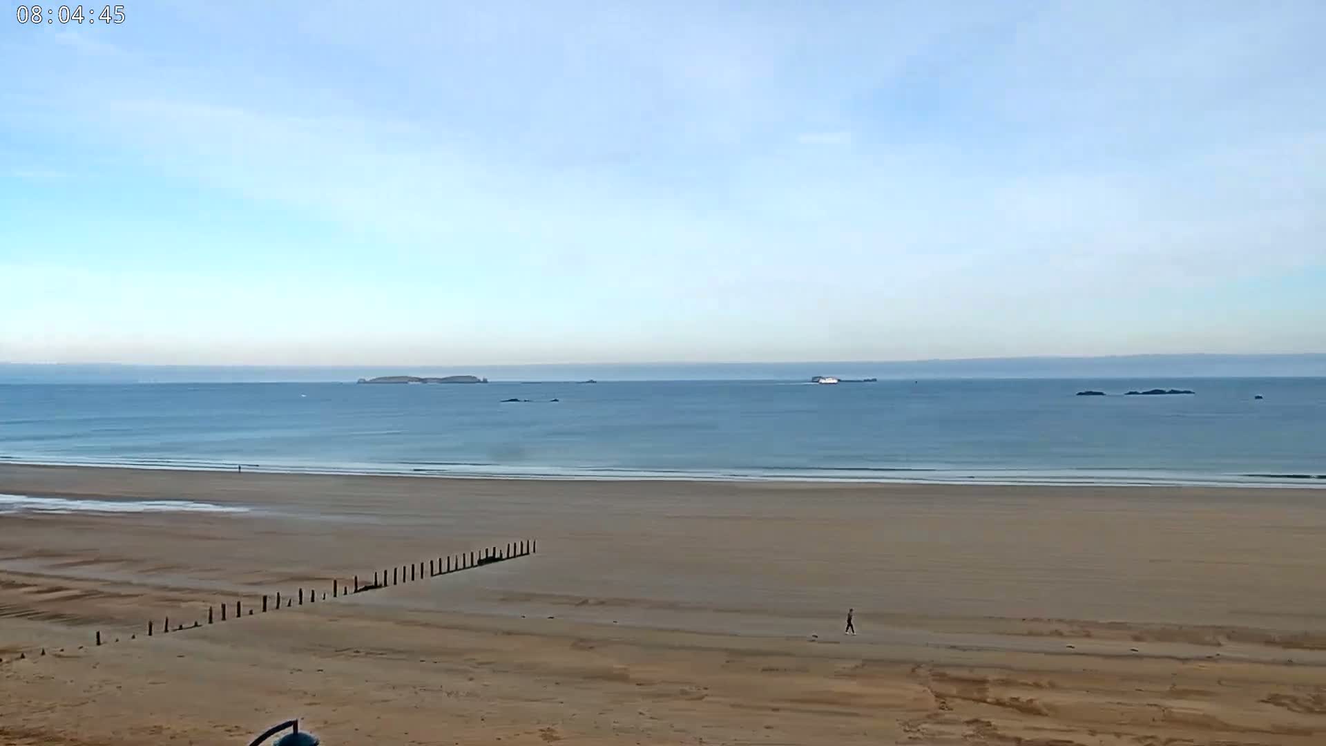 A person walks on a wide, sandy beach under a clear, light-blue sky, with calm ocean waters and distant islands visible on the horizon.