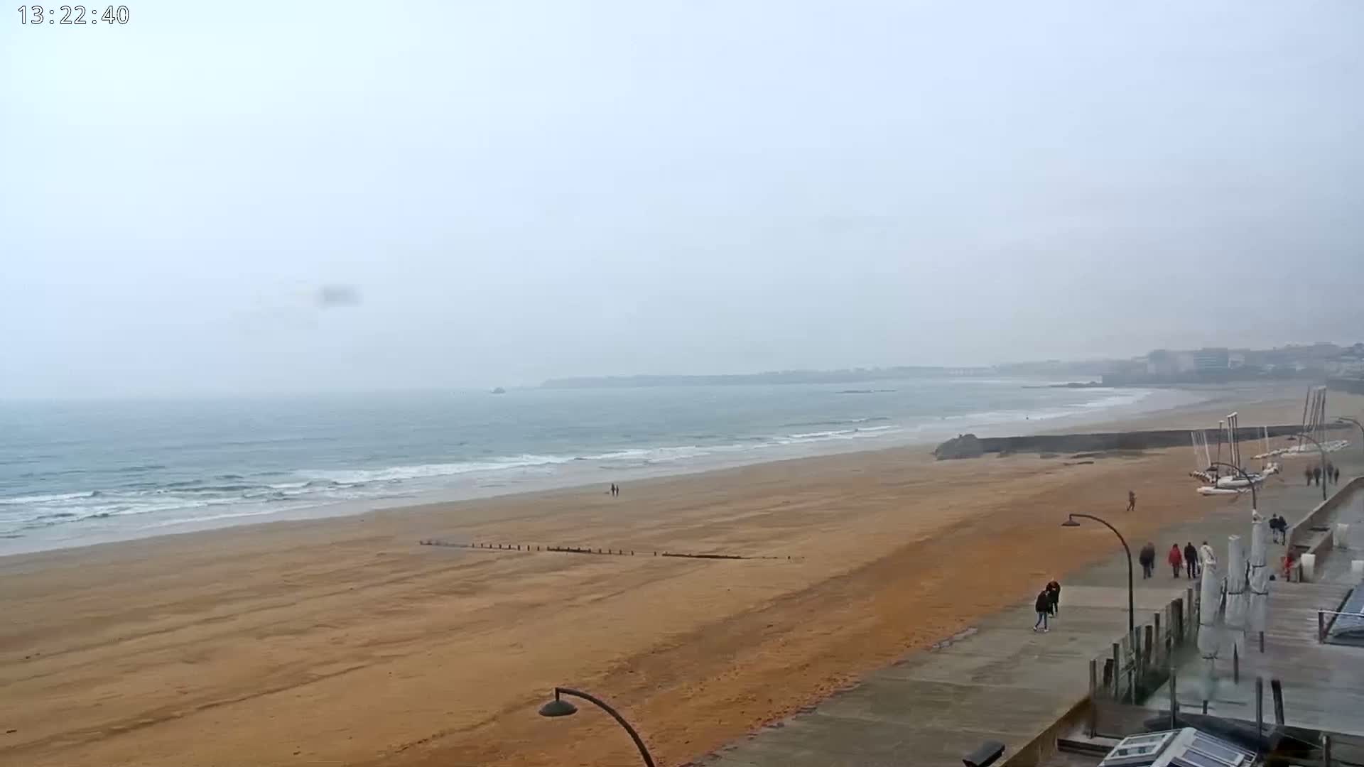 A wide sandy beach with gentle waves and a hazy overcast sky is viewed from a vantage point above a paved walkway with several people walking.