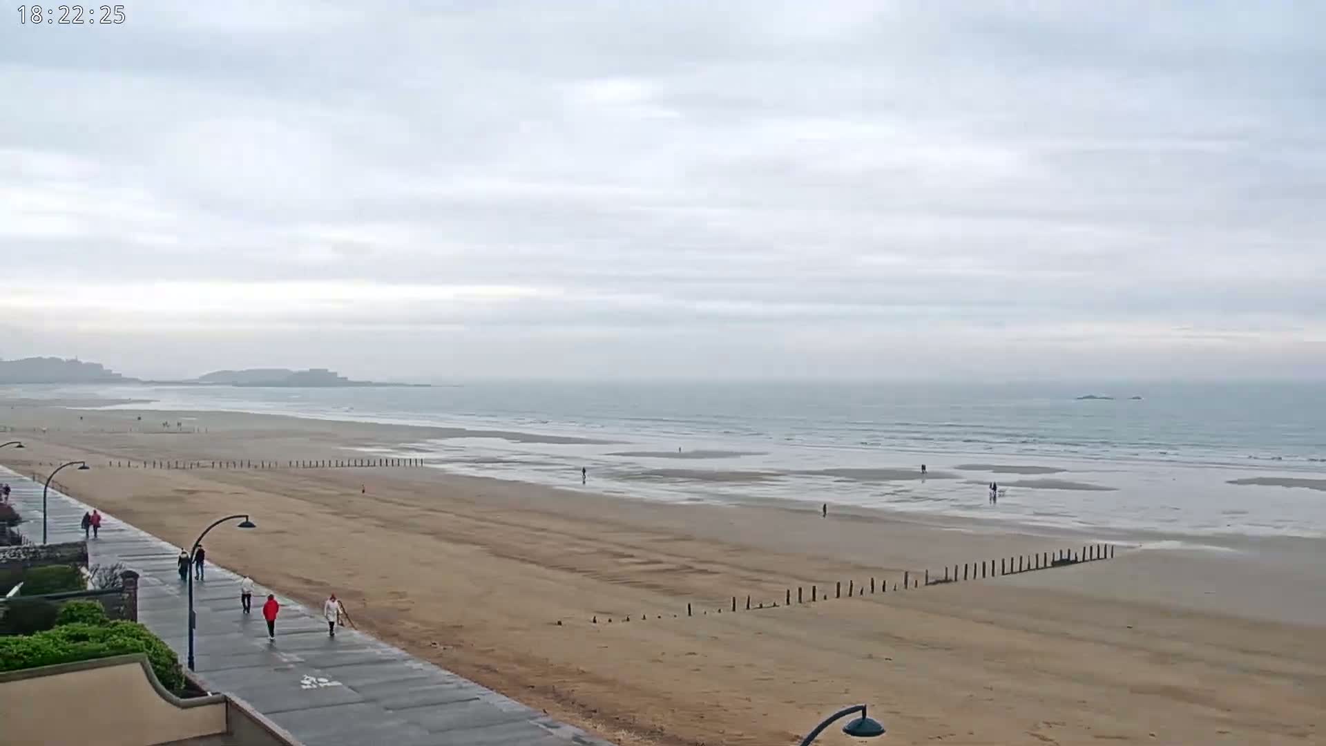 A long, sandy beach stretches to a calm, gray ocean under an overcast sky, with a few people walking on both the beach and a paved walkway.