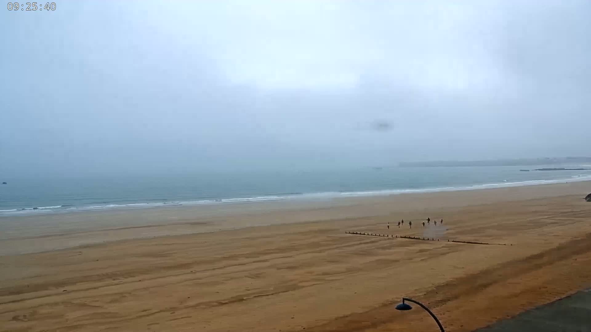A wide shot of a mostly empty sandy beach on an overcast day, with a few figures visible in the distance running along the water's edge.