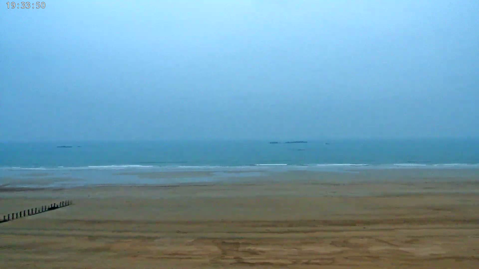 A wide sandy beach stretches to a calm, hazy ocean with a few small objects visible in the distance.