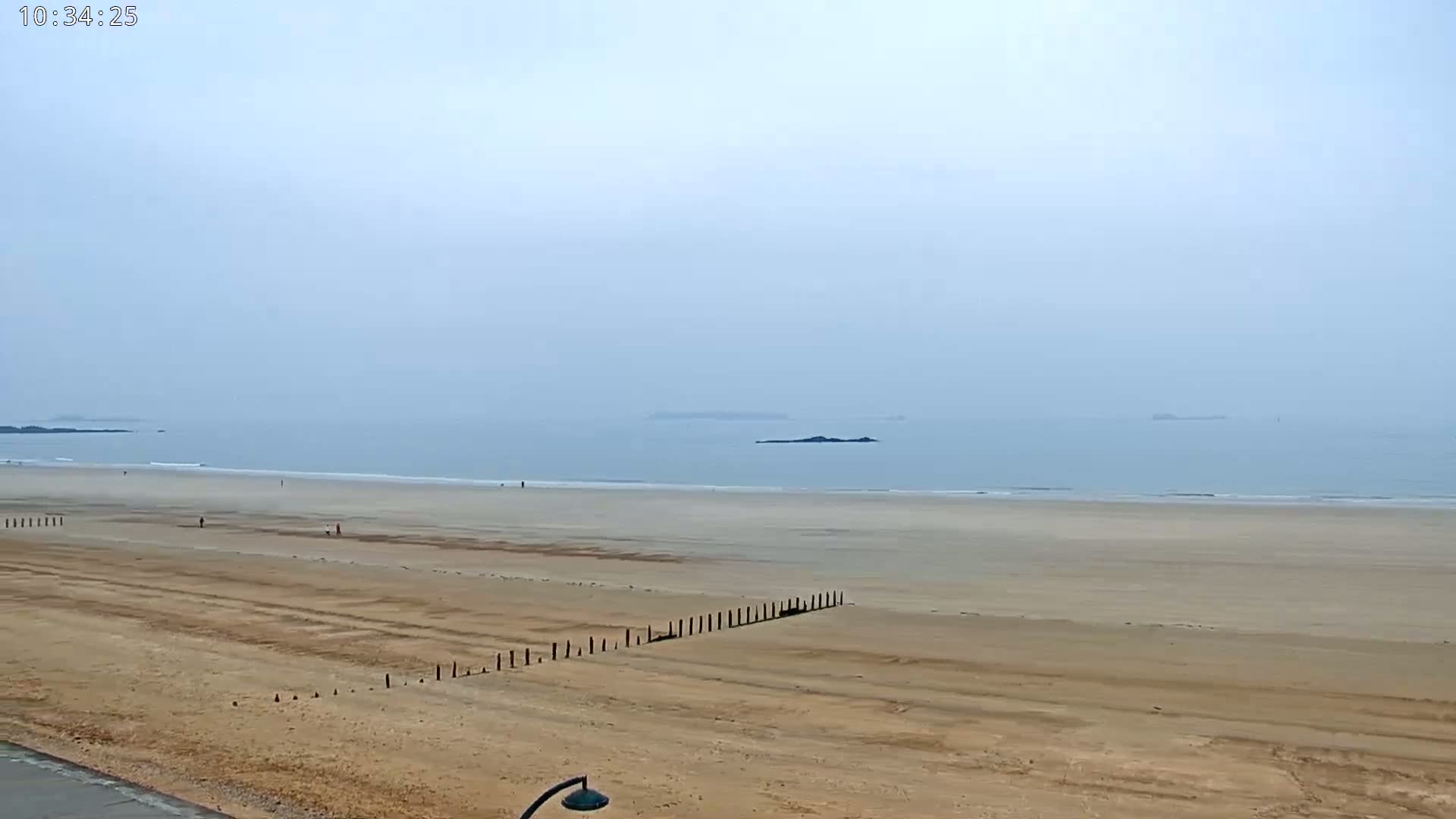 A hazy, overcast day reveals a wide expanse of sandy beach meeting a calm ocean with a few distant islands visible on the horizon.
