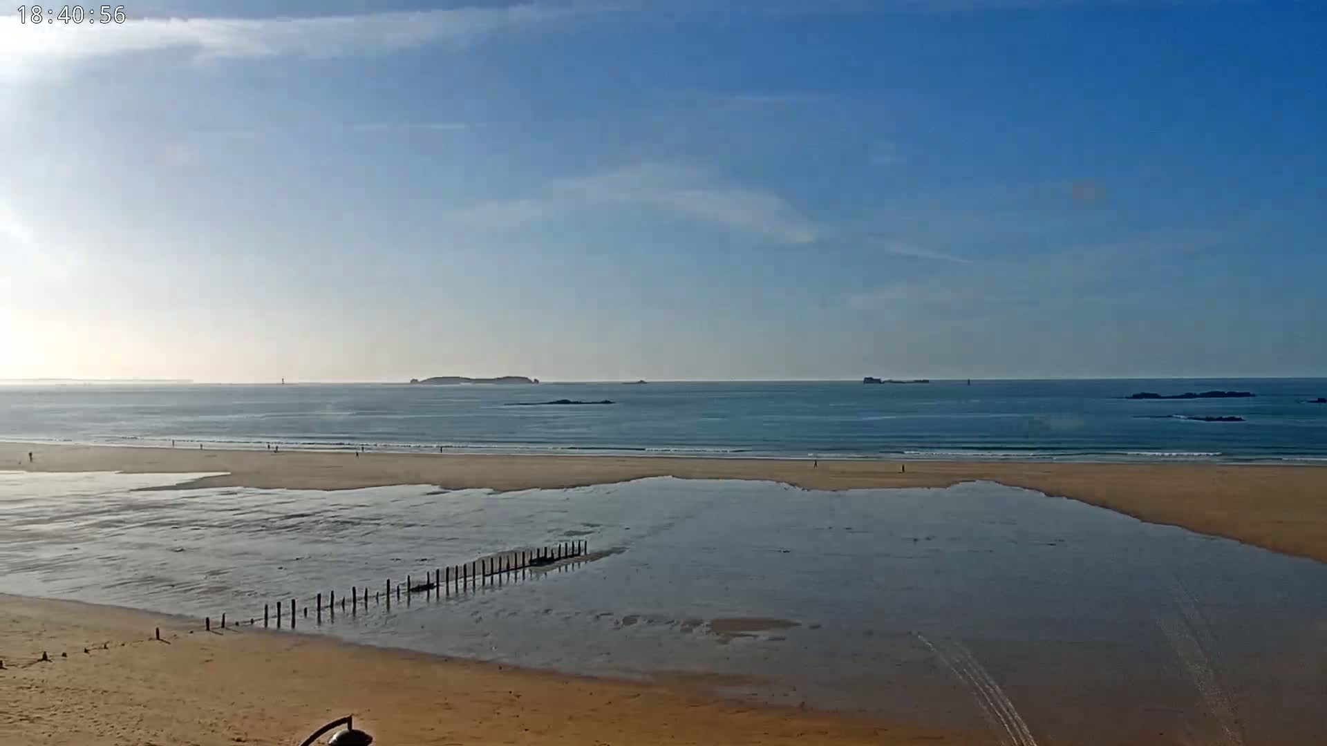 A sandy beach with calm ocean waves under a mostly clear blue sky shows a few people walking on the shore and a low wooden structure in the shallow water.