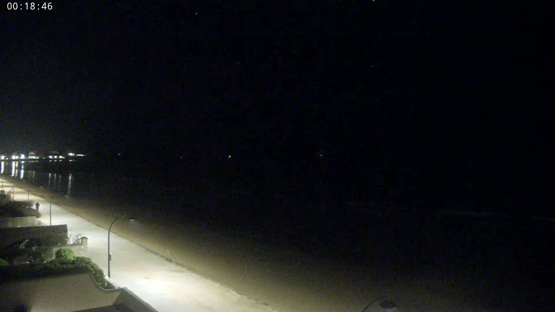 A nighttime view of a mostly empty beach and a dimly lit road next to it, under a clear, starry sky.