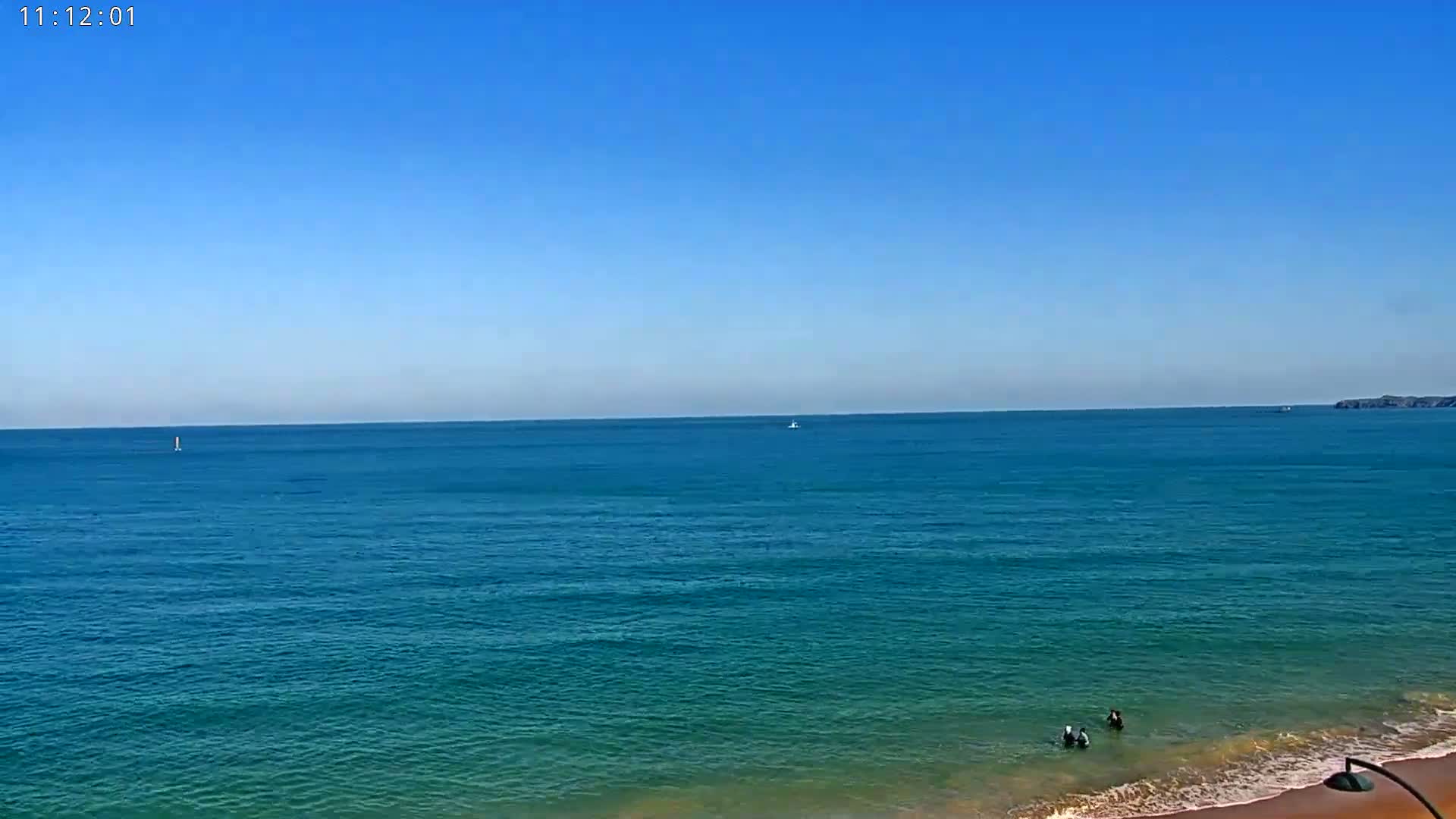 A calm, blue ocean under a clear sky meets a sandy beach where a few people are wading in the shallow water.