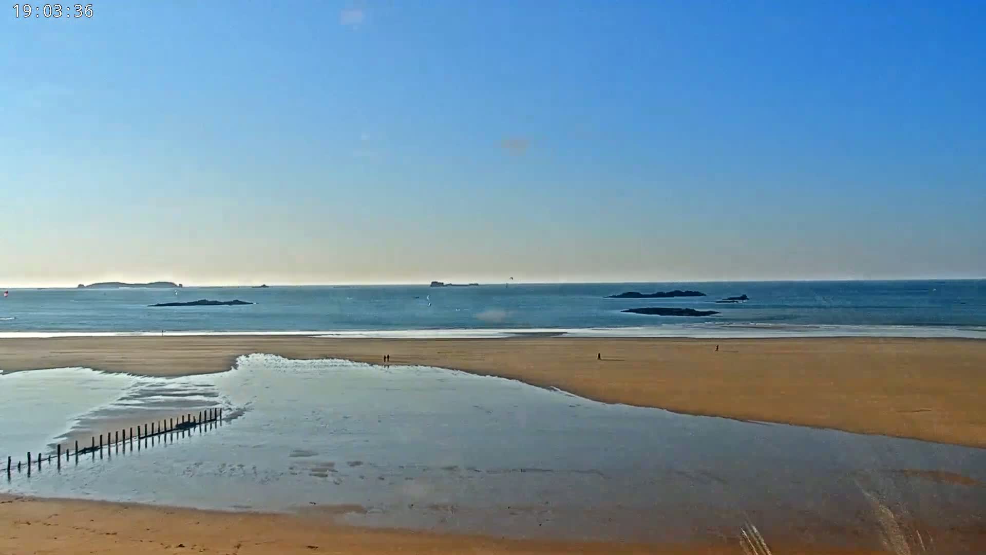 A sandy beach stretches to a calm ocean under a clear blue sky, with a few small islands visible in the distance and a wooden post fence partially submerged in shallow water.