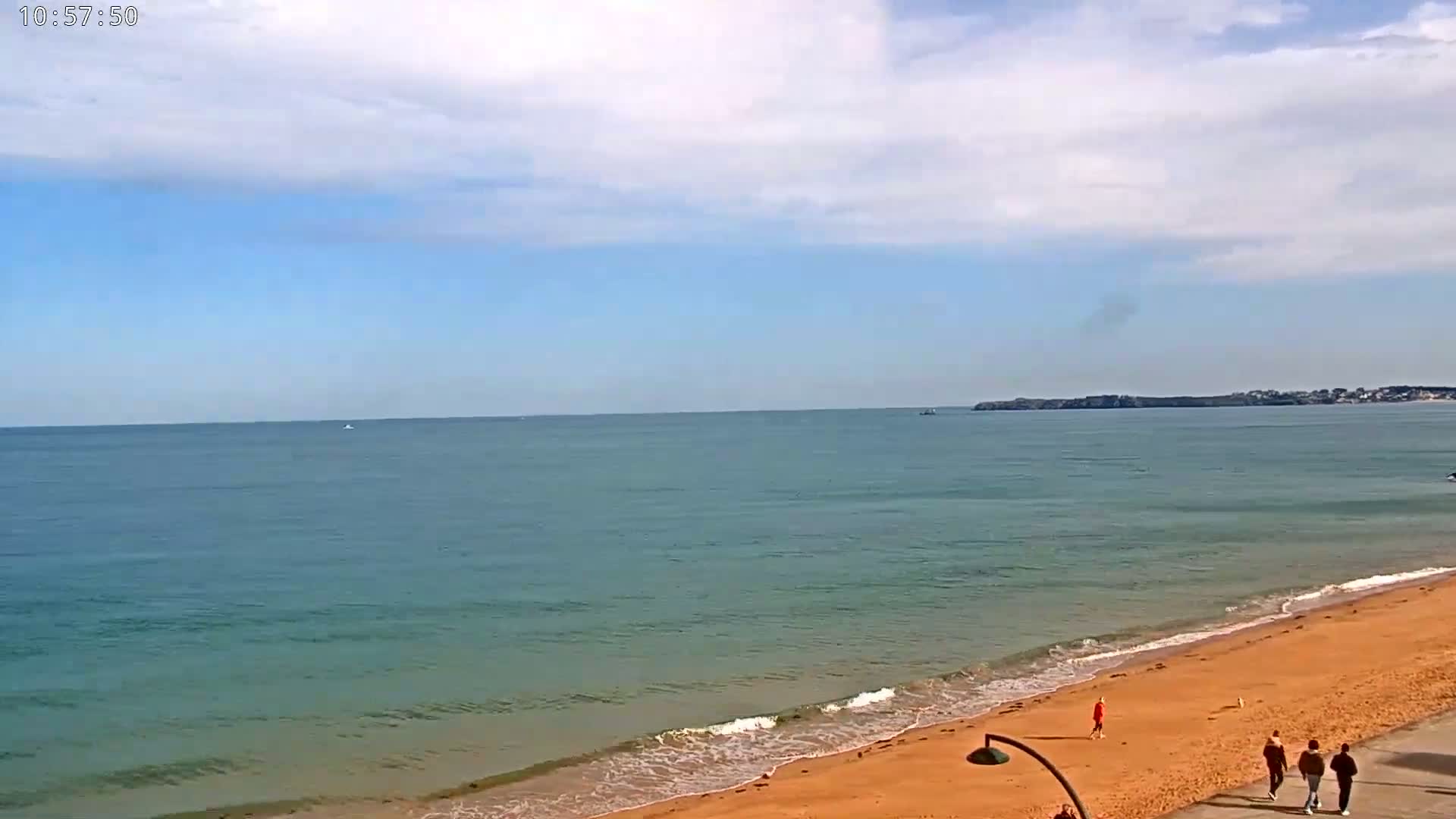 A sandy beach with a calm, teal ocean under a partly cloudy blue sky, a few people walk along the shore.