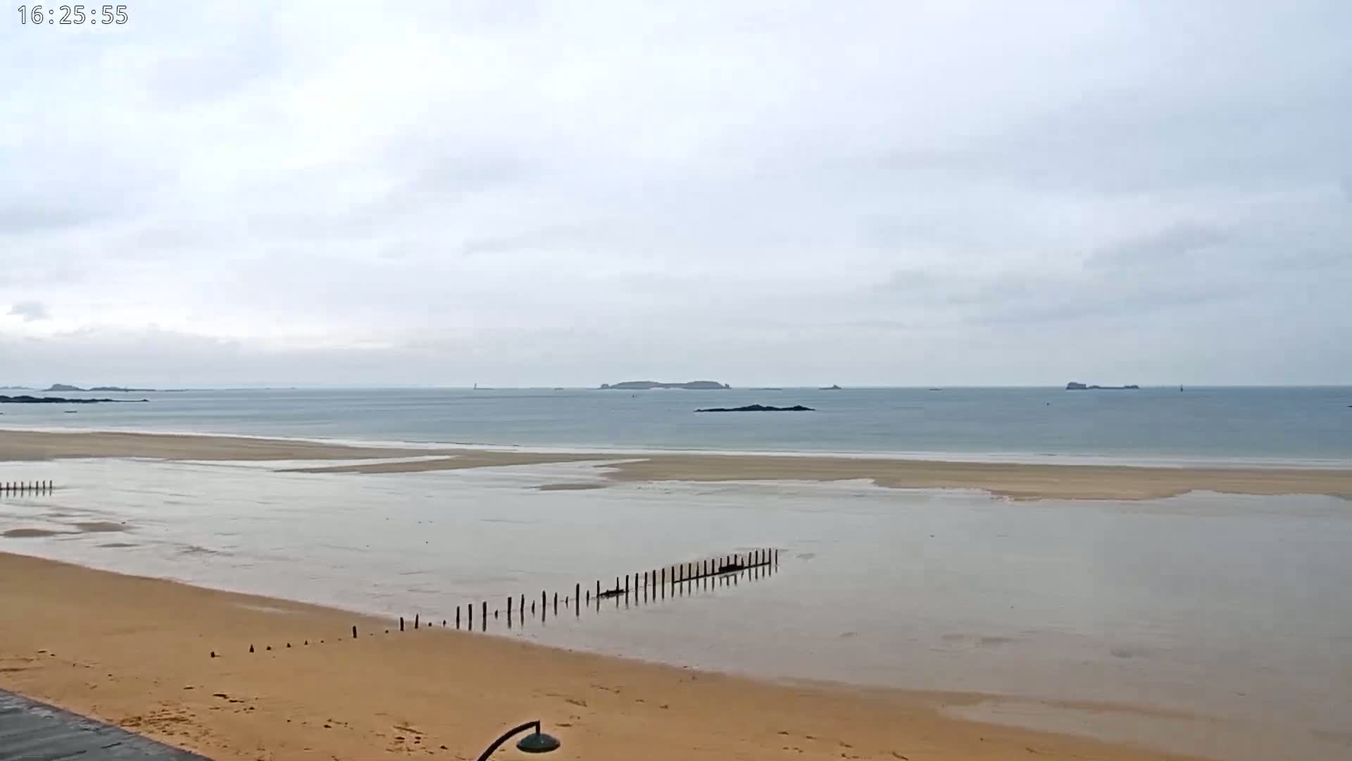 A wide sandy beach with shallow, calm water stretches to a calm sea under an overcast sky.