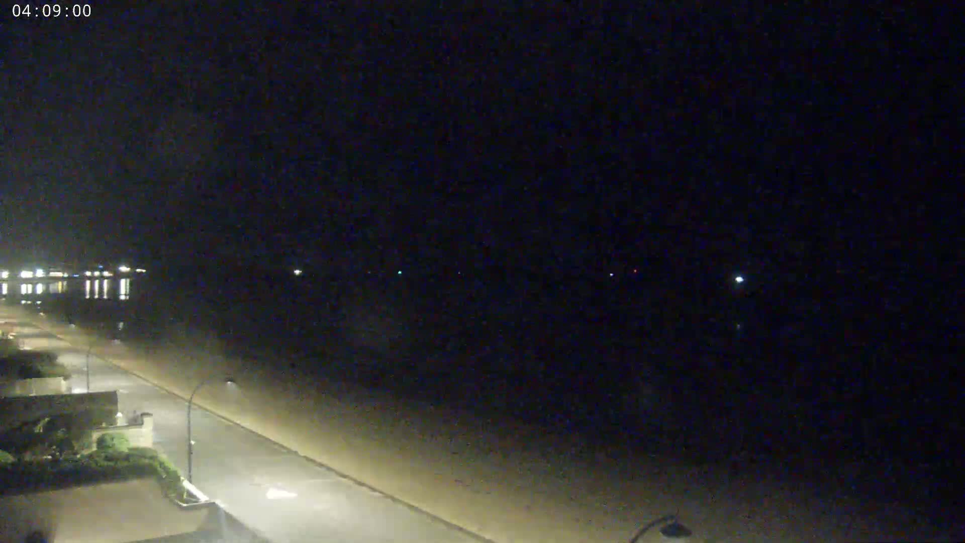 A dark, nighttime beach scene shows a mostly empty road next to a shoreline under a clear sky.