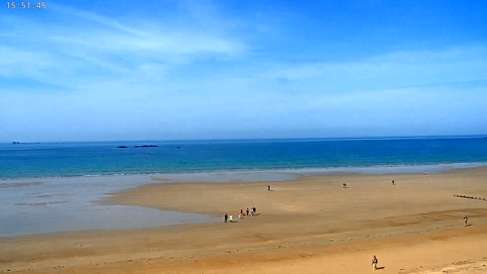 A wide sandy beach stretches to a calm, blue ocean under a mostly clear, bright blue sky.
