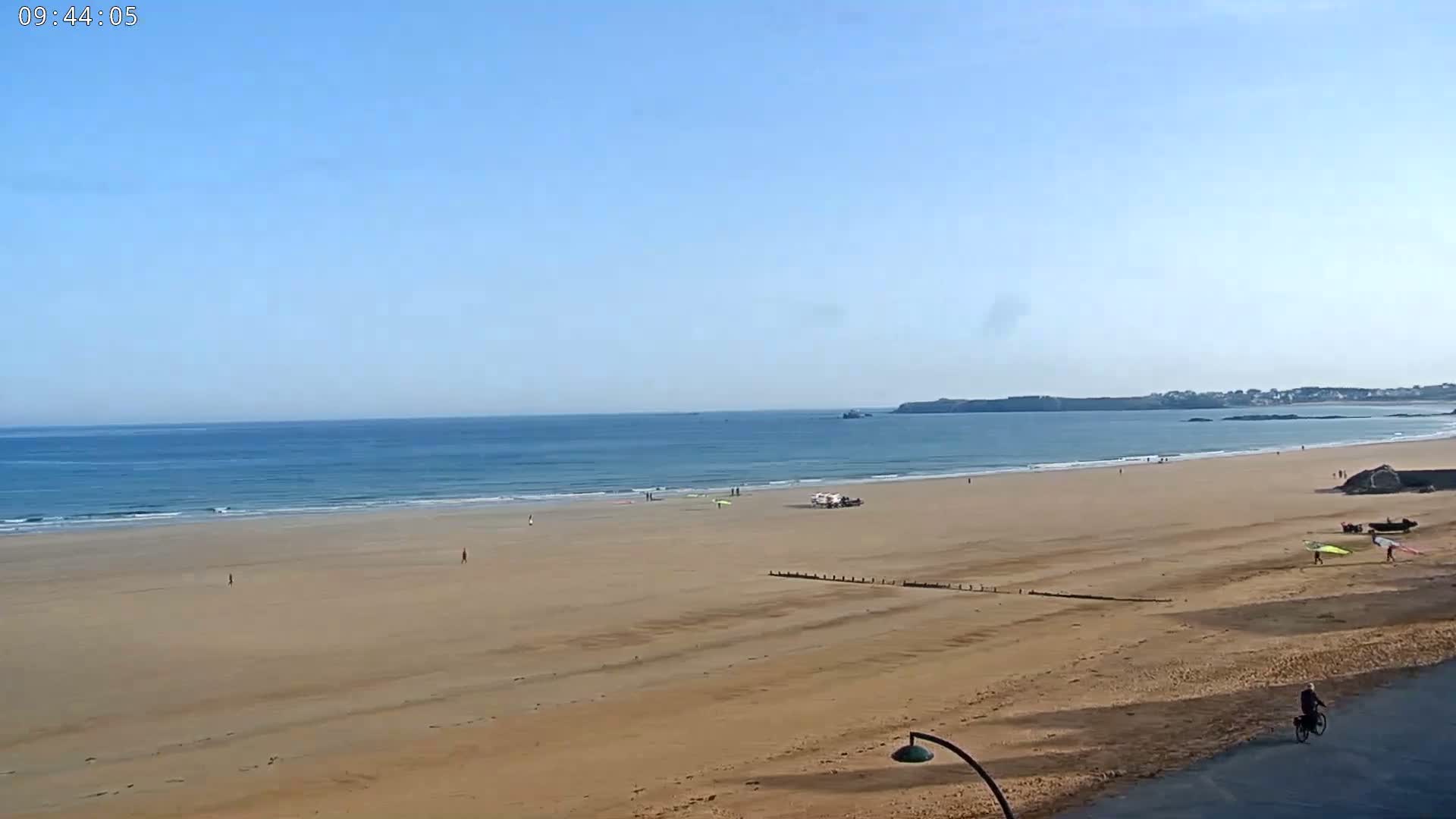 A sandy beach stretches out to a calm ocean under a clear blue sky, with a few people and windsurfing equipment visible.