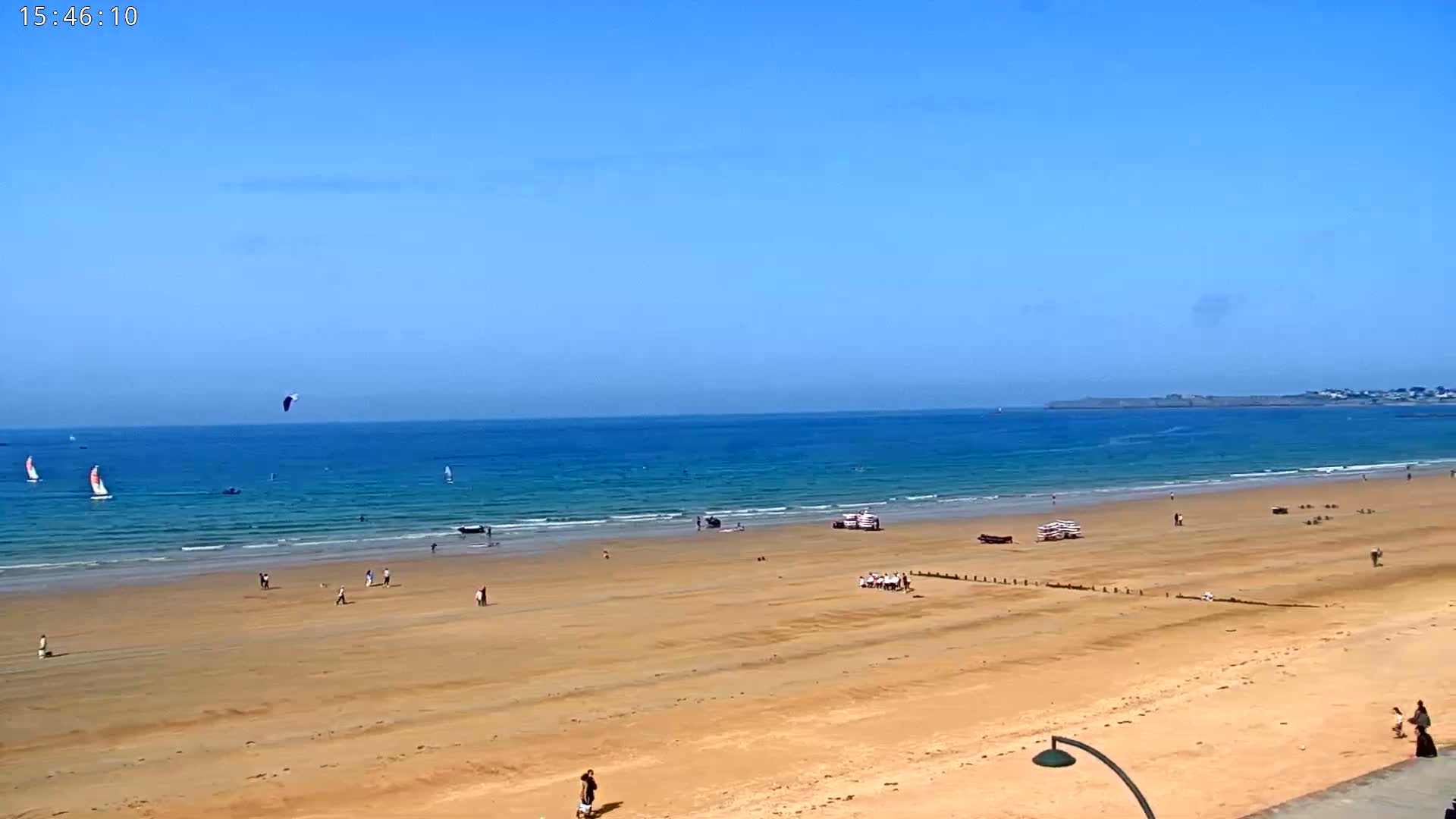 A wide sandy beach stretches to a calm, blue ocean under a clear blue sky, with several people scattered across the sand and a few sailboats and a kite surfer visible in the water.
