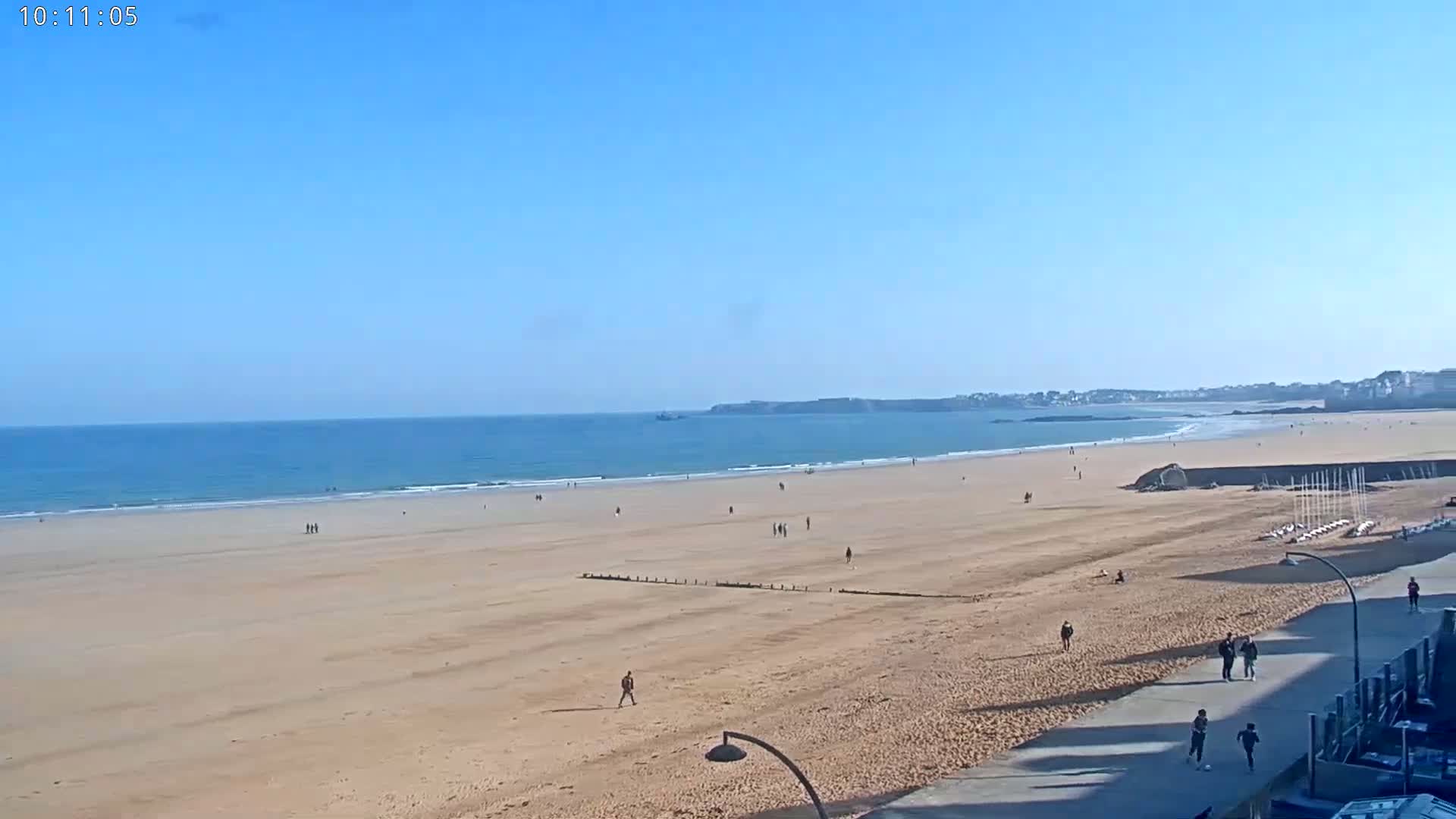 A wide sandy beach stretches to a calm blue ocean under a clear blue sky, with a few people scattered across the sand and a paved walkway along the right edge.