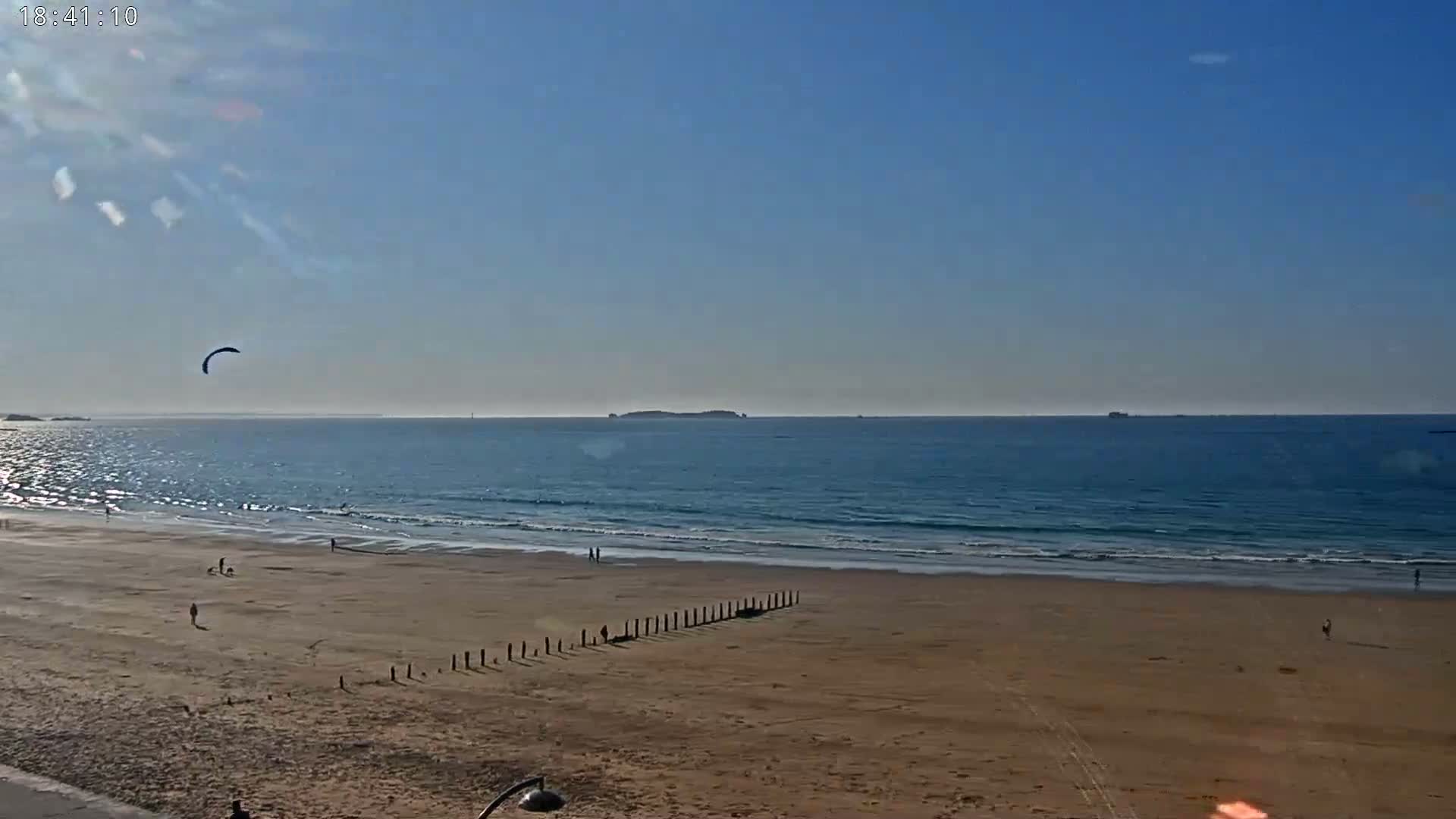 A sandy beach with a few people and a kite surfer, under a mostly sunny sky, overlooks a calm ocean with islands in the distance.