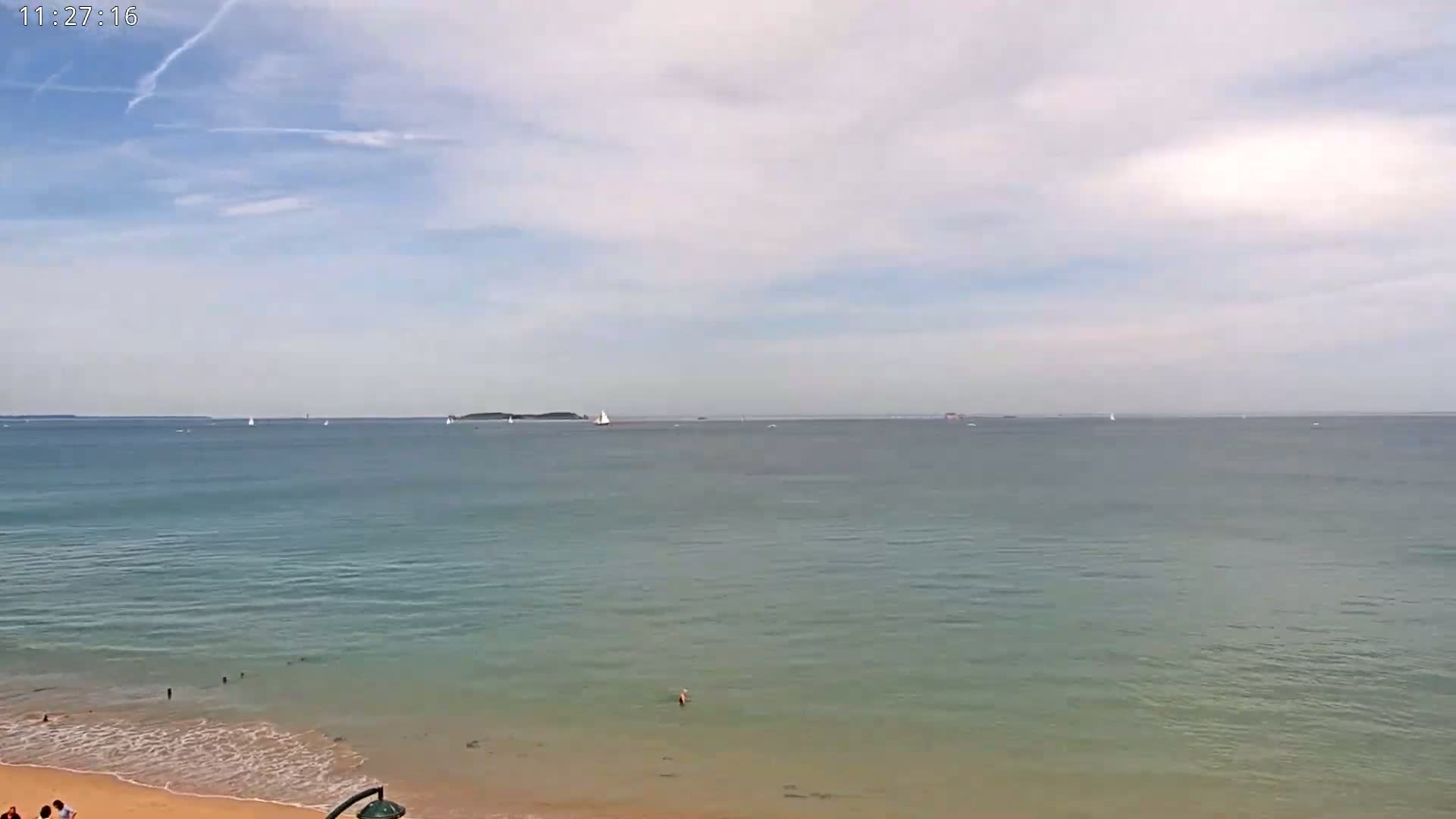 A sandy beach meets a calm, light-blue ocean under a mostly cloudy sky, with several sailboats visible on the water.