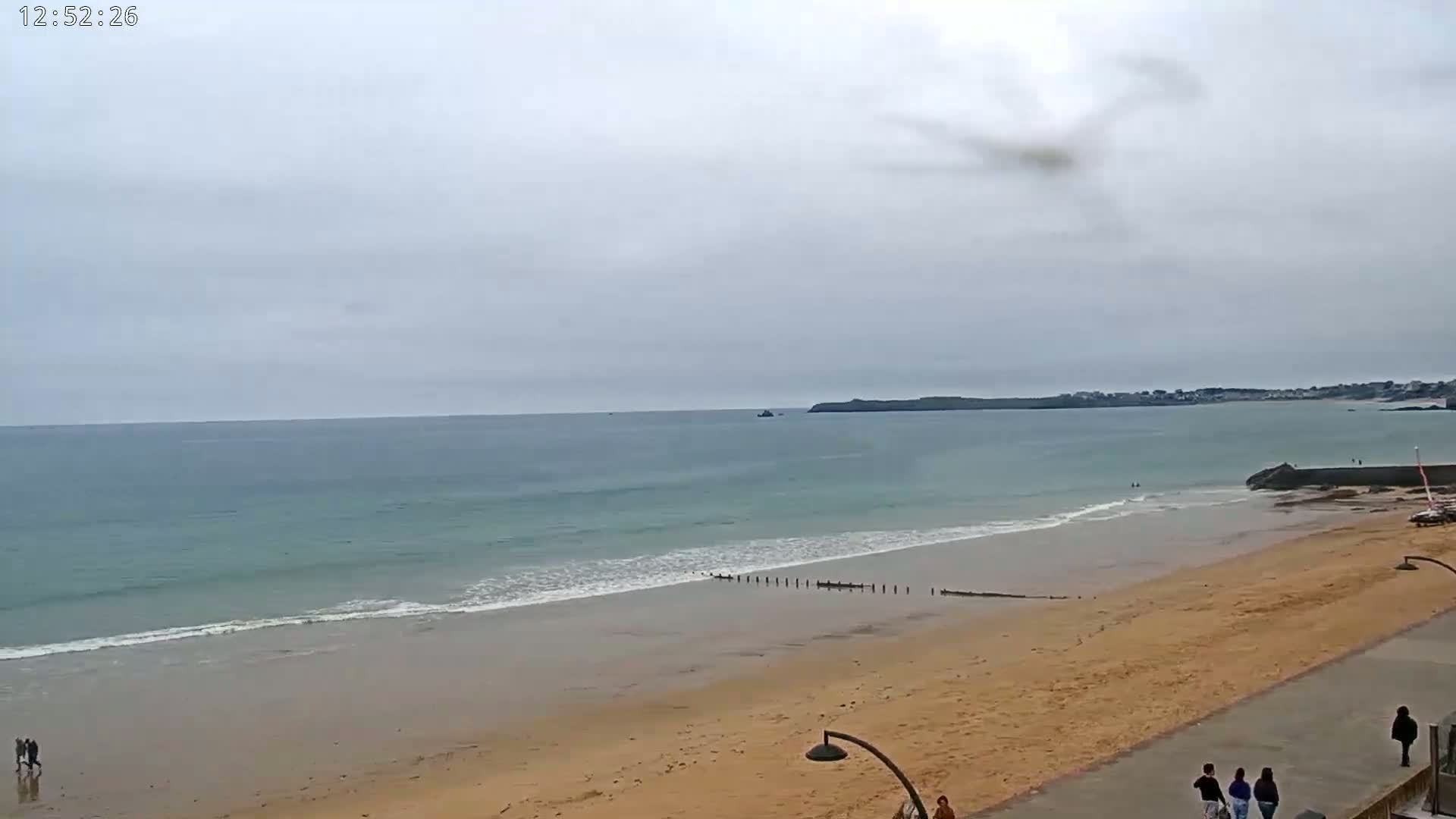 A sandy beach with gentle waves under an overcast sky, and a few people walking along the shore.