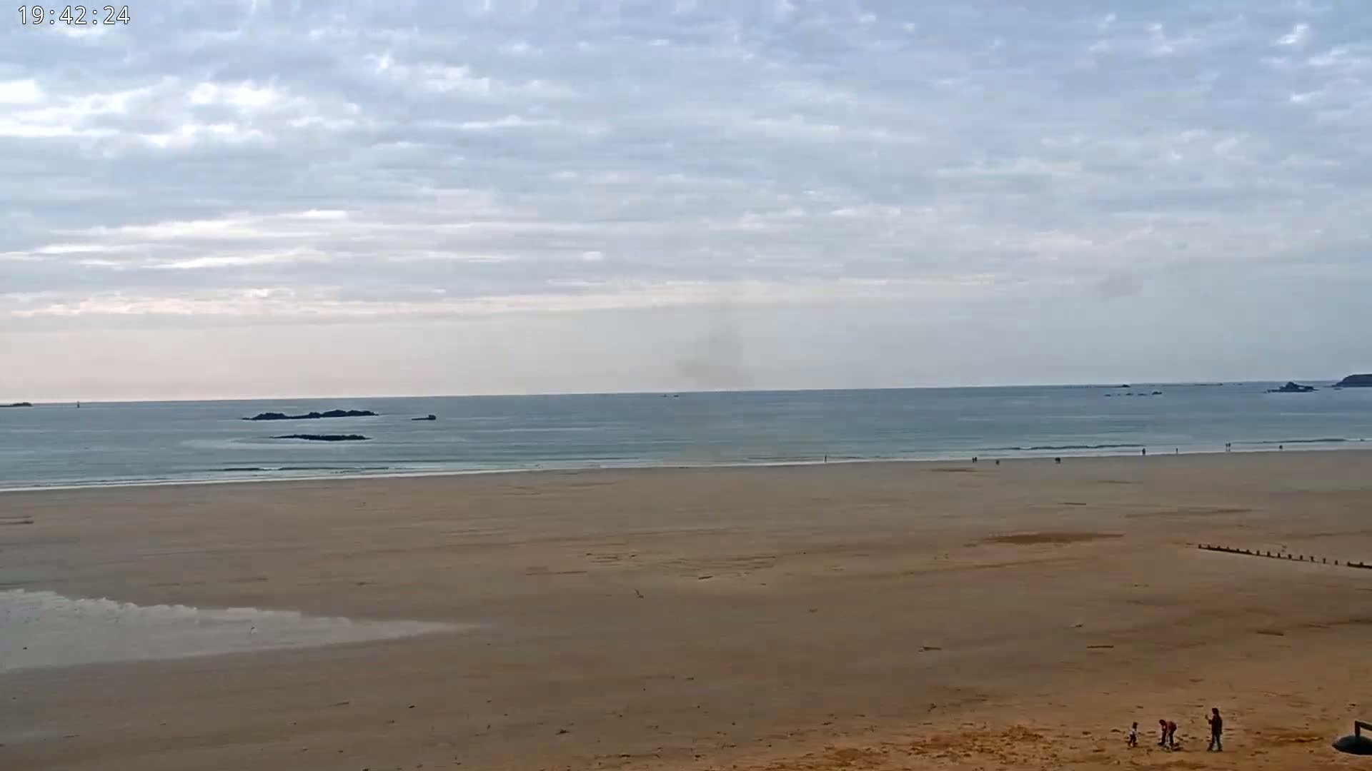 A wide sandy beach stretches to a calm ocean under a mostly cloudy sky, with a few people visible on the sand and small islands in the distance.