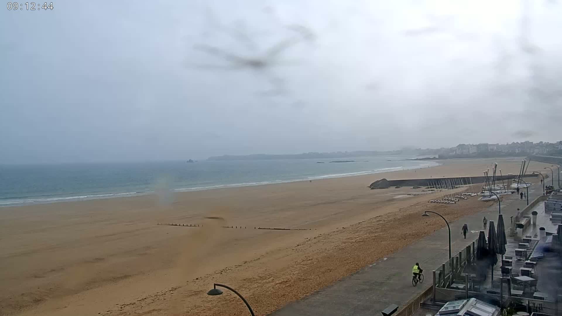A wide, sandy beach stretches along a calm ocean under an overcast sky, with a person cycling along a paved walkway near the shoreline and a few people walking near some stored sailboats.