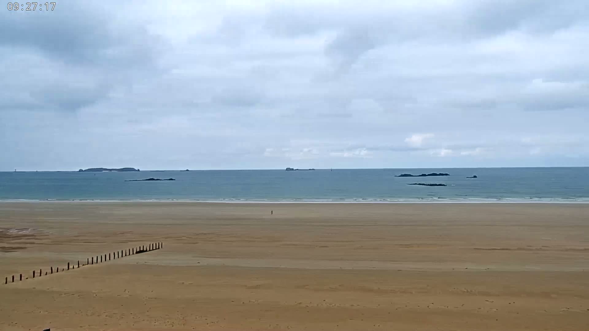 A long, sandy beach stretches to a calm, light-blue ocean under a mostly cloudy sky, with several small islands visible in the distance.