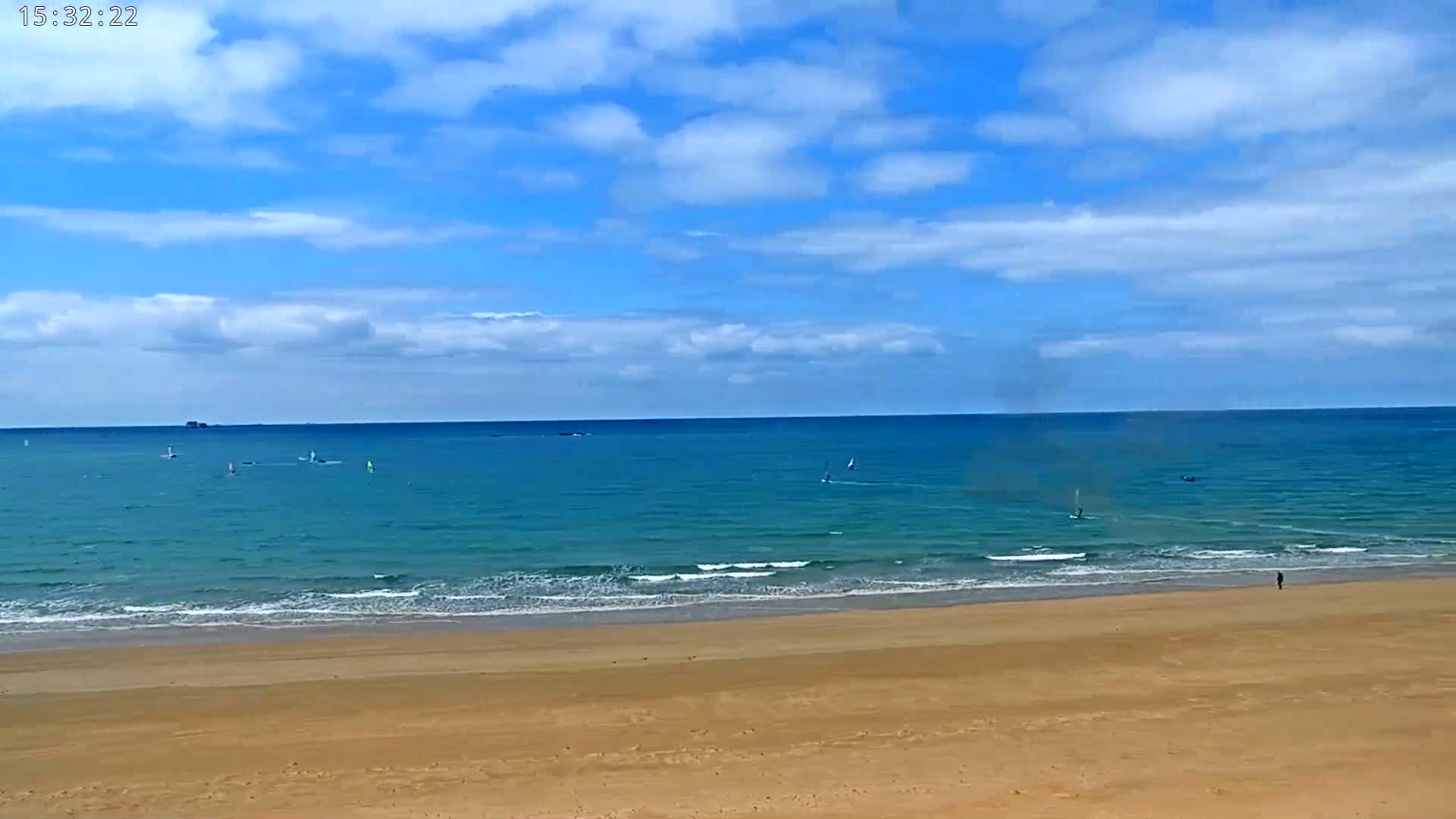A sandy beach stretches out to a calm teal ocean under a mostly sunny sky with scattered clouds, where several windsurfers are visible in the distance.