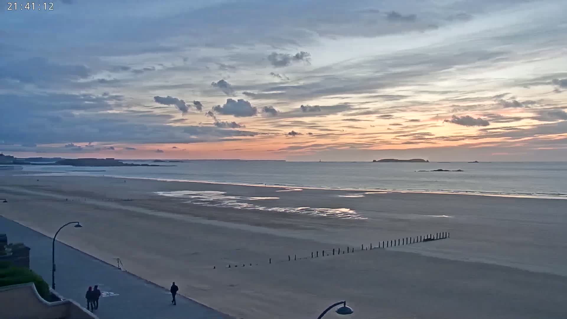 A wide sandy beach stretches to a calm ocean under a pastel sunset sky with scattered clouds.