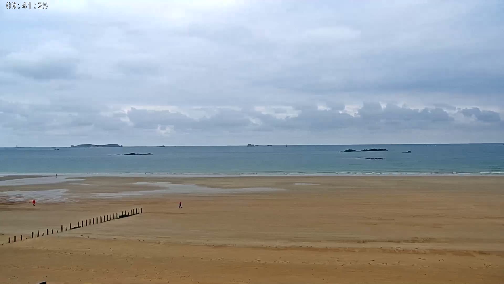A wide expanse of sandy beach stretches to a calm sea under a cloudy sky, with small islands visible in the distance.