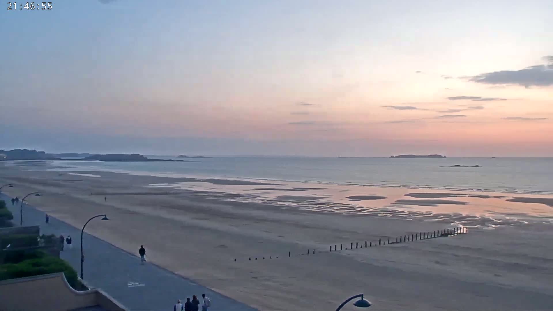 A wide sandy beach stretches to a calm ocean under a pale pink and blue sunset sky, with a few people walking along a paved path next to it.