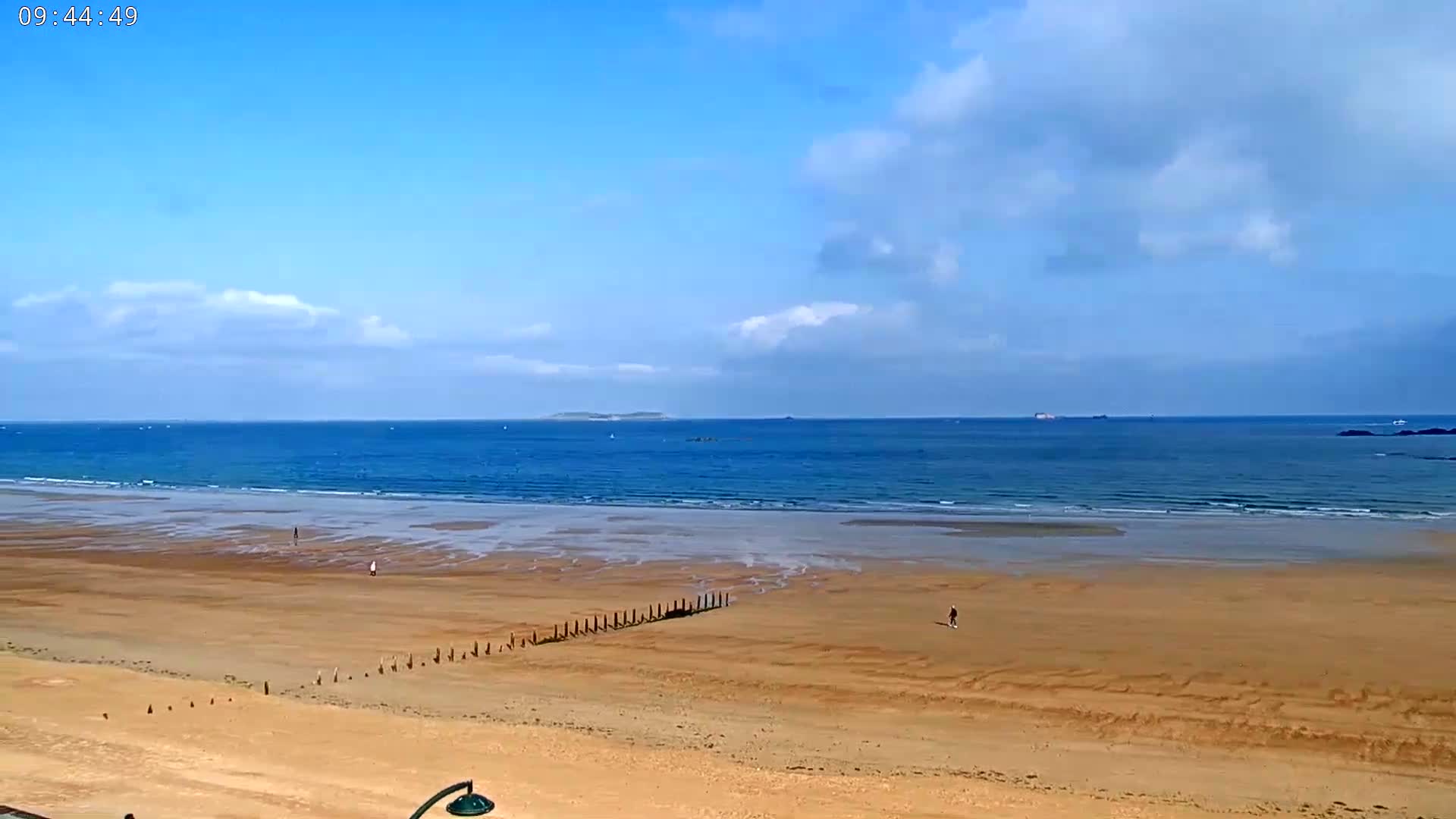 A mostly sunny day at a wide, sandy beach with calm ocean waves and a distant island.