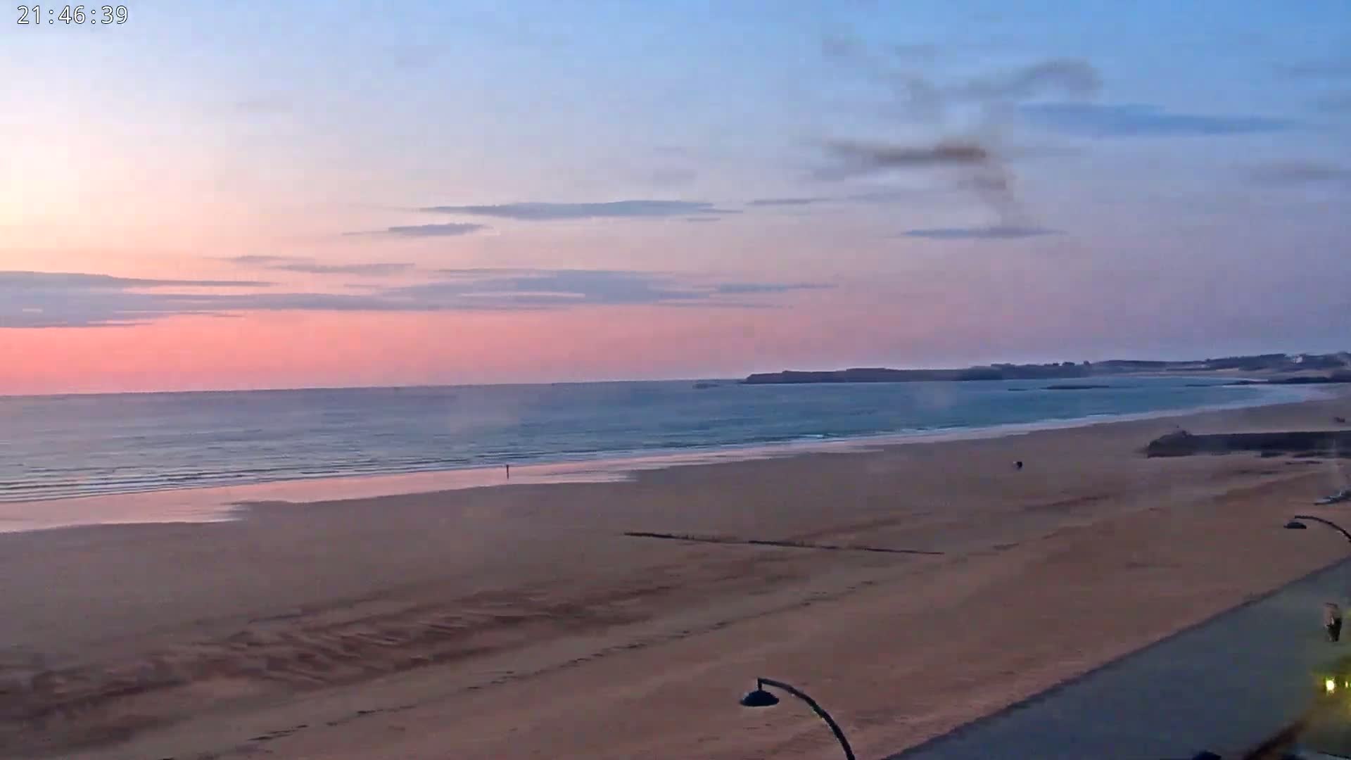 A pink and blue sunset over a calm ocean and sandy beach, with a few people visible on the shore.