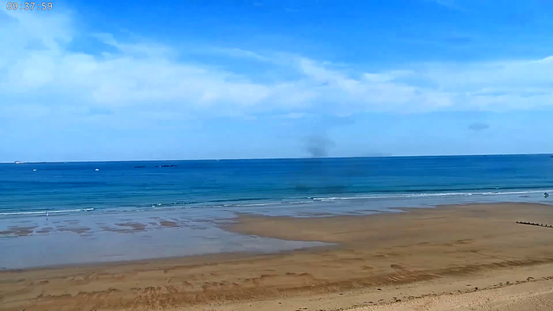A sandy beach stretches out to a calm, blue ocean under a mostly clear sky with a few scattered clouds.