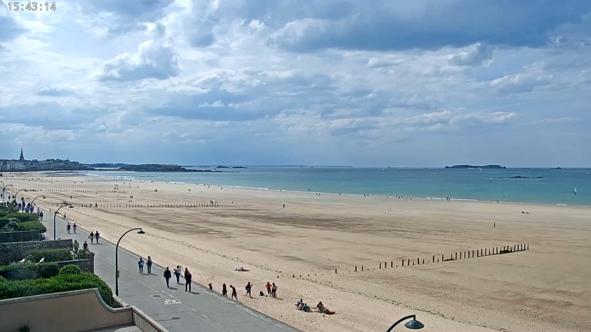 A wide sandy beach stretches to a calm, teal ocean under a partly cloudy sky, with people walking along a paved path and relaxing on the sand.