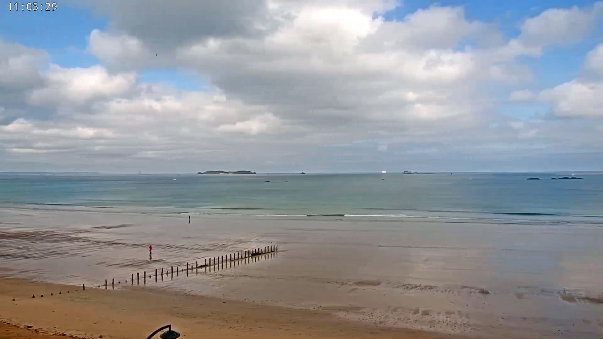 A partly cloudy sky overlooks a wide sandy beach with calm, teal-colored ocean waters and distant islands.