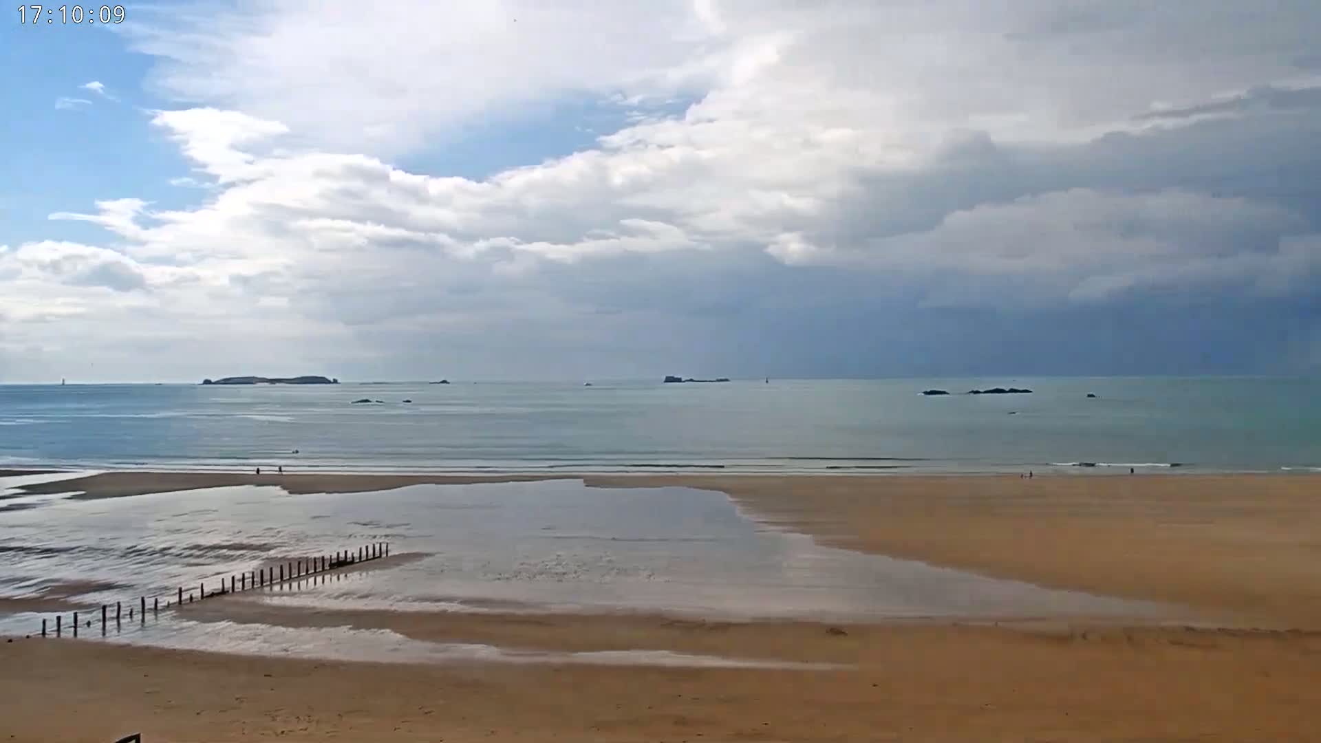 A sandy beach with a partially receding tide, a wooden breakwater, and several small islands visible on the horizon under a partly cloudy sky.