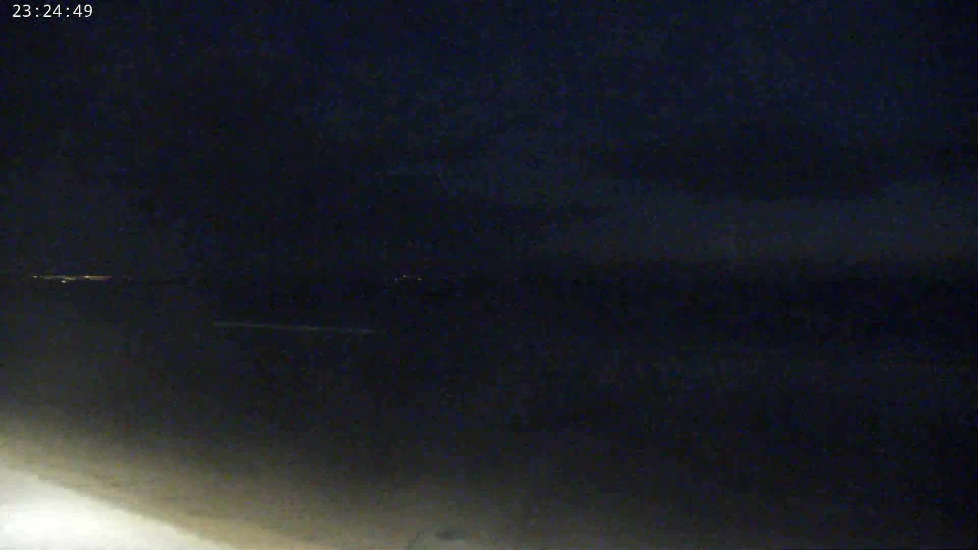 A dark nighttime scene shows a body of water with a distant shoreline of lights under a mostly clear sky.