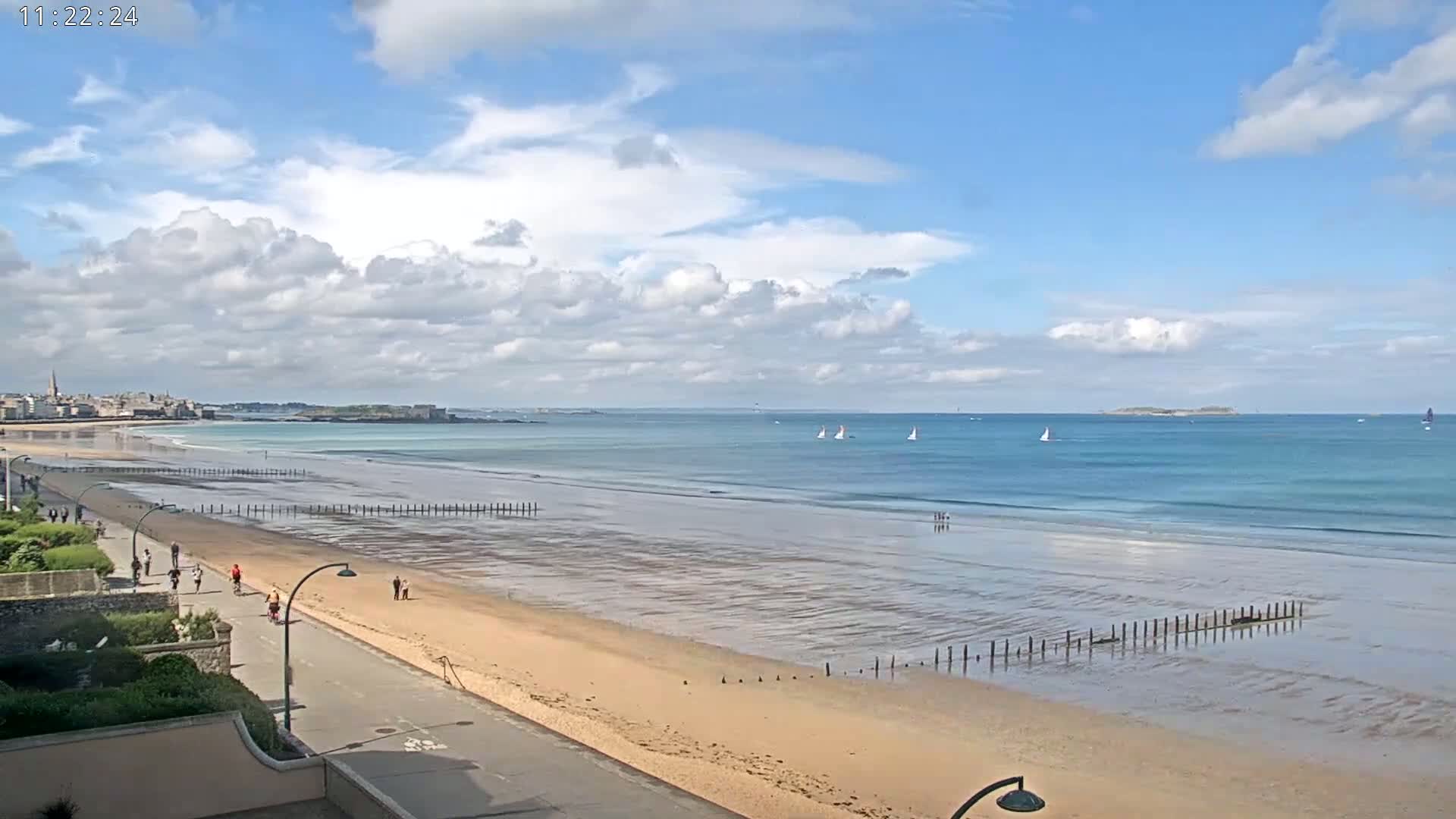 A long sandy beach with a paved walkway and people walking and cycling, stretches toward a calm ocean under a partly cloudy sky with several sailboats visible in the distance.