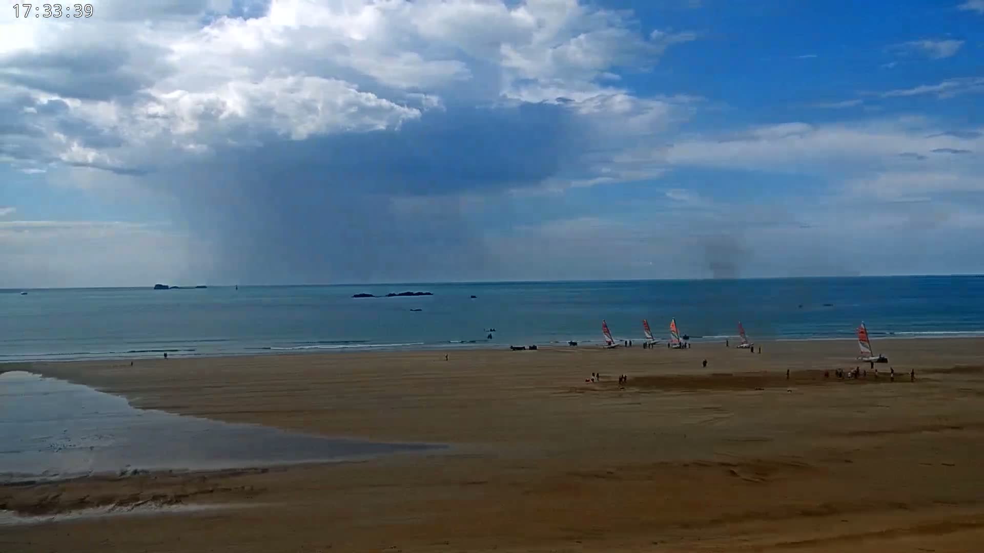 A sandy beach with several small sailboats on the shore is visible under a partly cloudy sky with rain falling over the ocean in the distance.