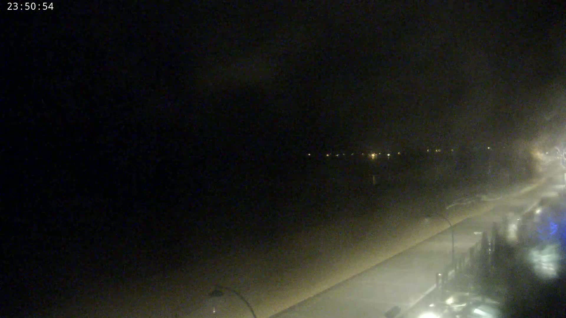 A dark, nighttime scene shows a beach and a roadway with faint lights in the distance, under hazy or overcast conditions.